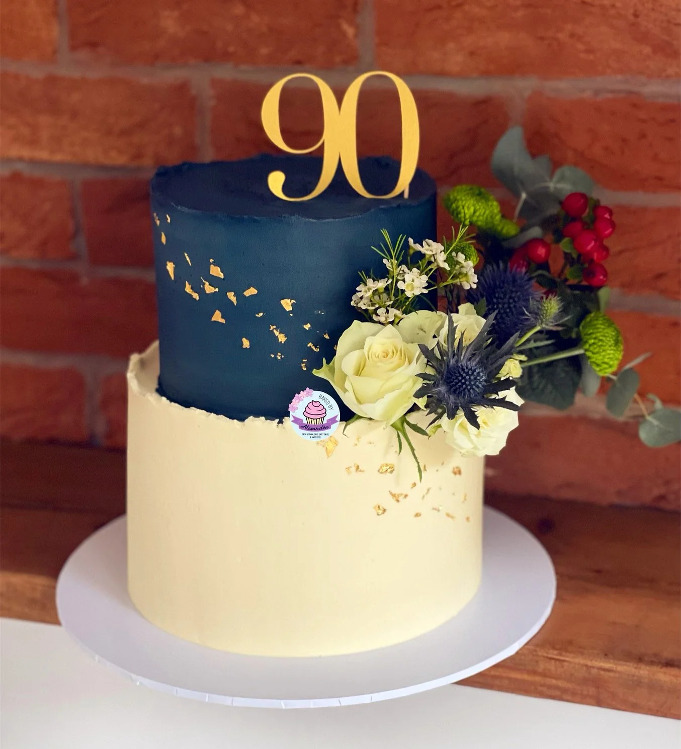 Two-tiered blue and white cake with "90" topper and floral decorations.