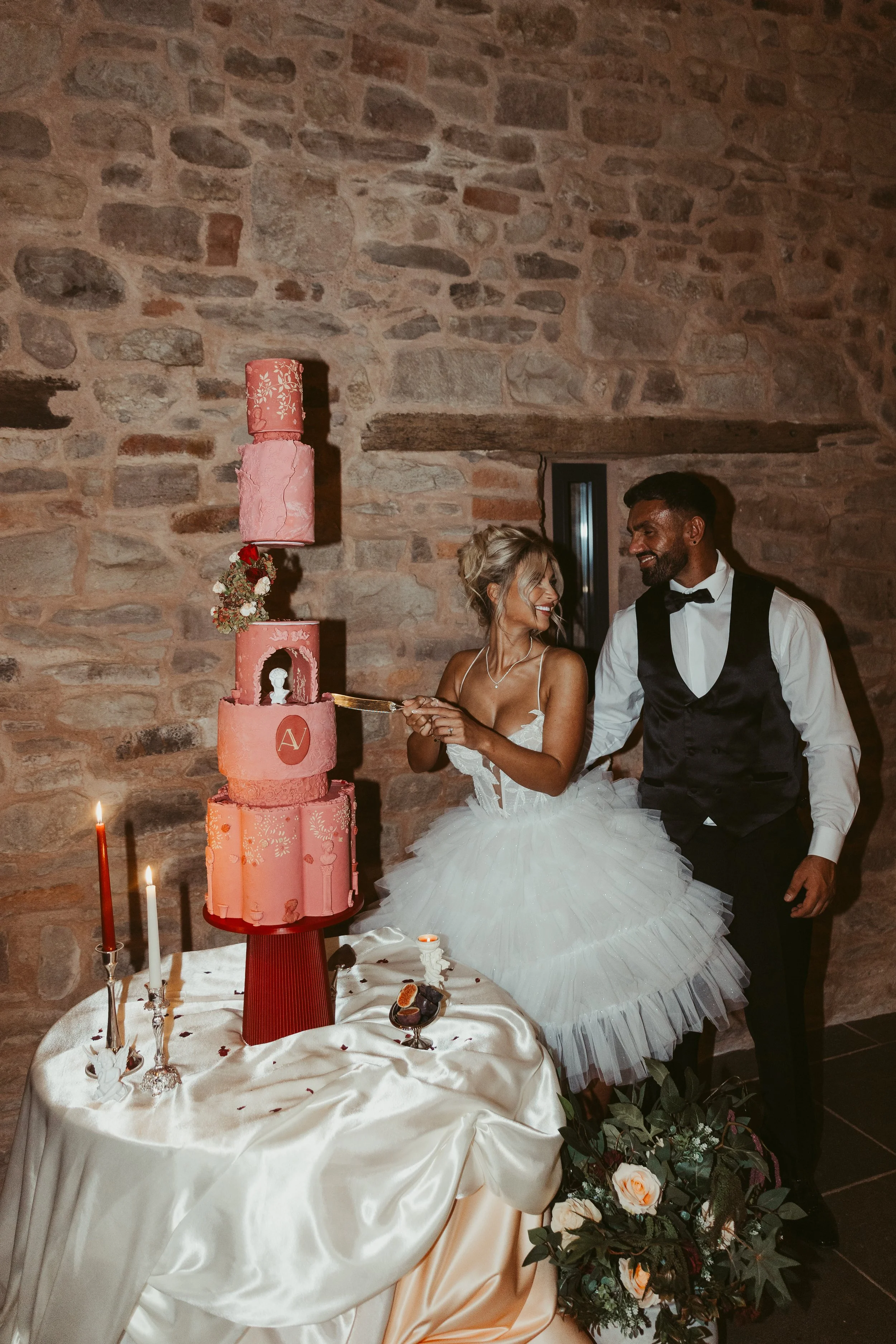 A bride and groom celebrating with a wedding cake topped with a pink and floral design, standing in front of a rustic stone wall.