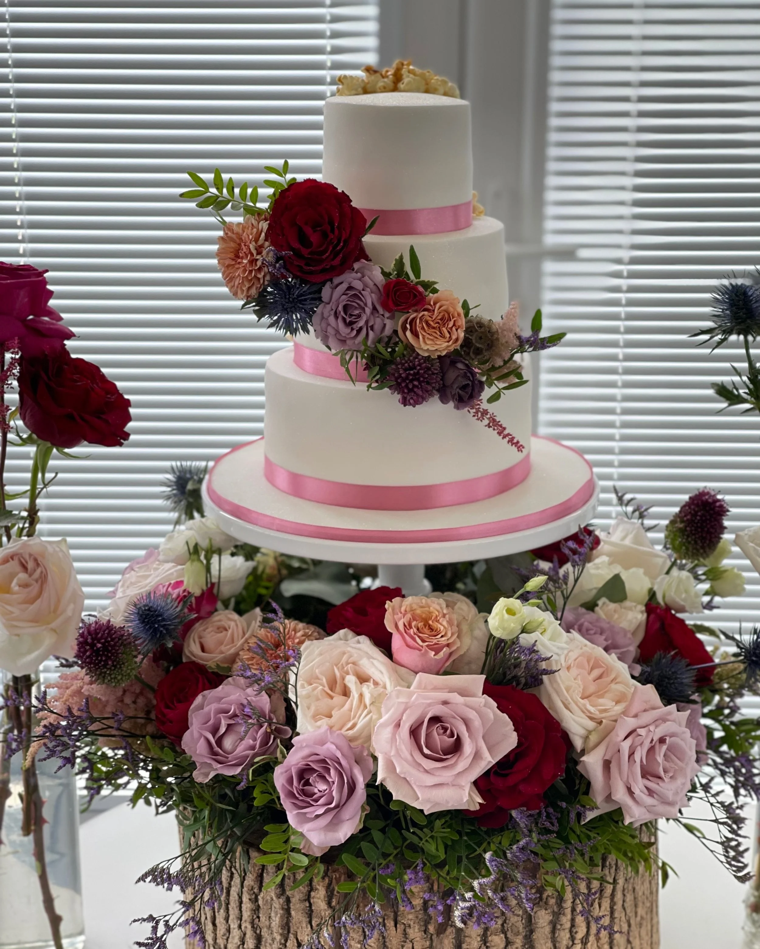 Three-tiered white wedding cake with pink ribbons and floral decorations, surrounded by a base of assorted roses and greenery.