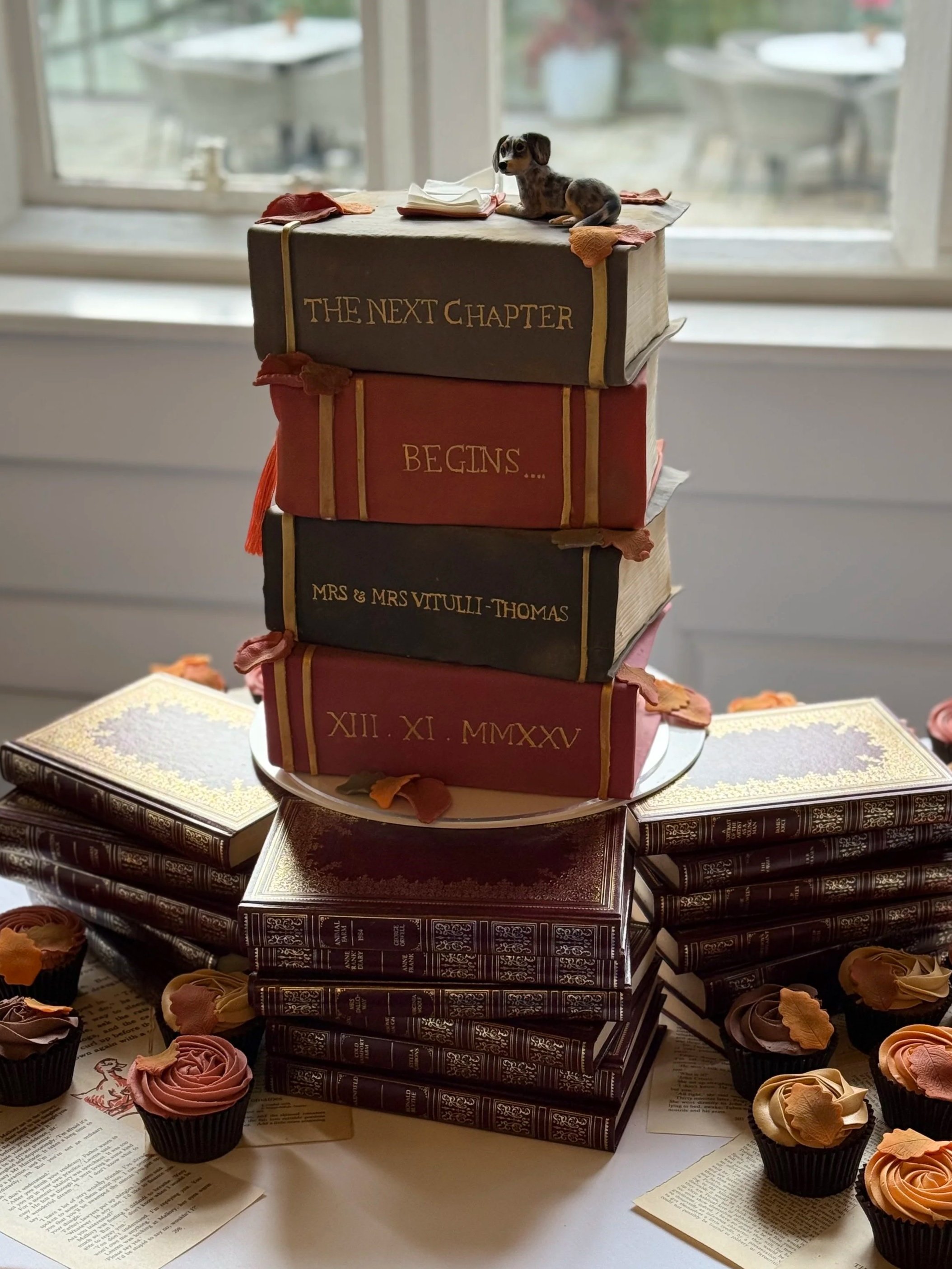 Four-tiered book wedding cake with autumnal colours and personalised hand painting. The cake is decorated with falling leaves nd a dog reading a book. 