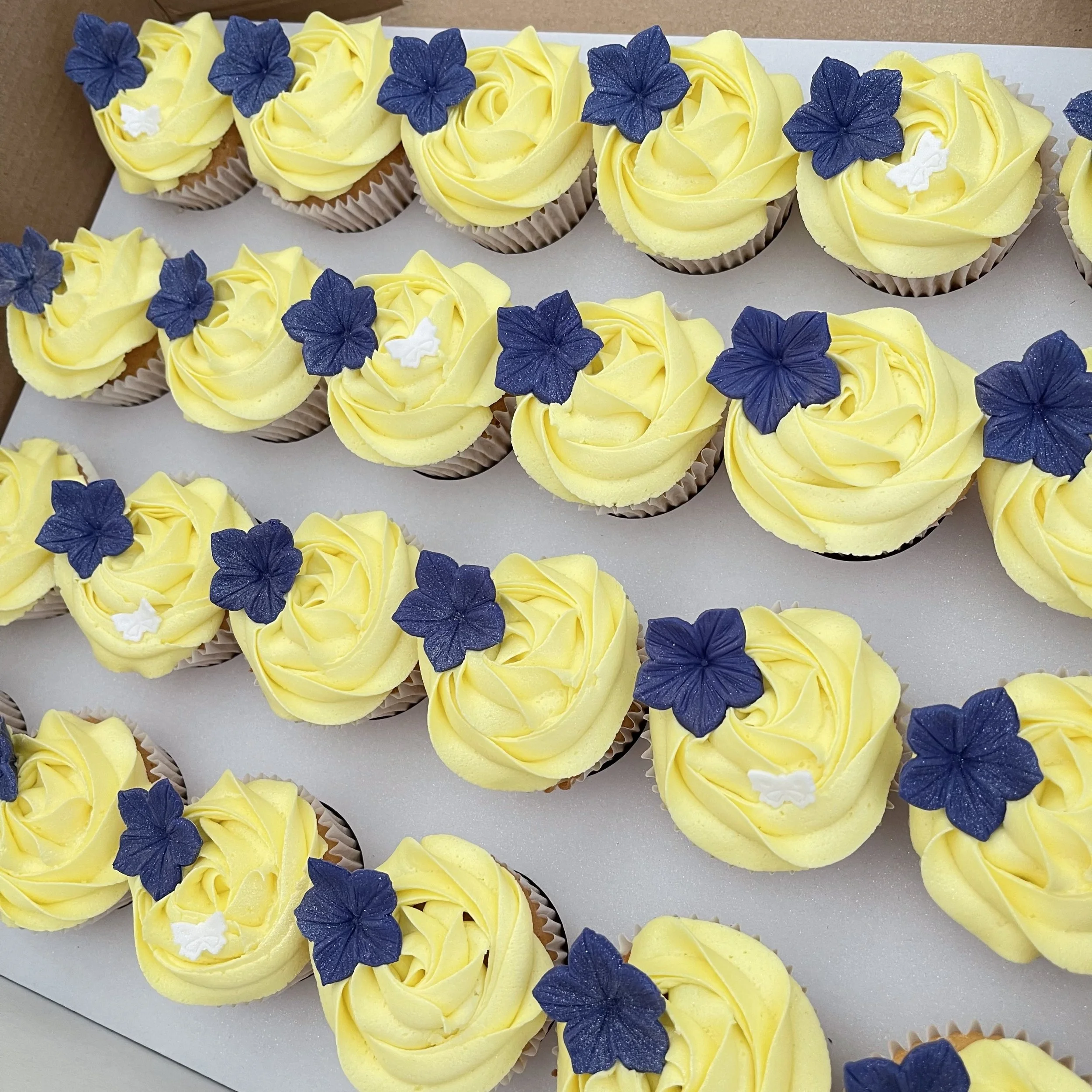 Yellow cupcakes with navy blue and white flower decorations arranged in rows.