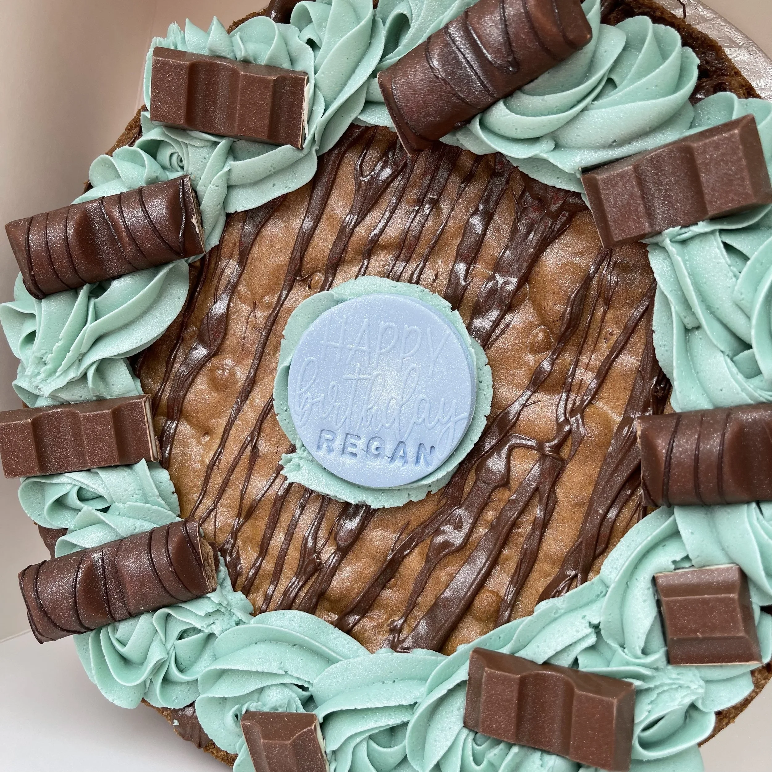 Chocolate chip cookie cake with chocolate drizzle, topped with mint green frosting swirls, chocolate bars, and a blue fondant circle with a birthday message.