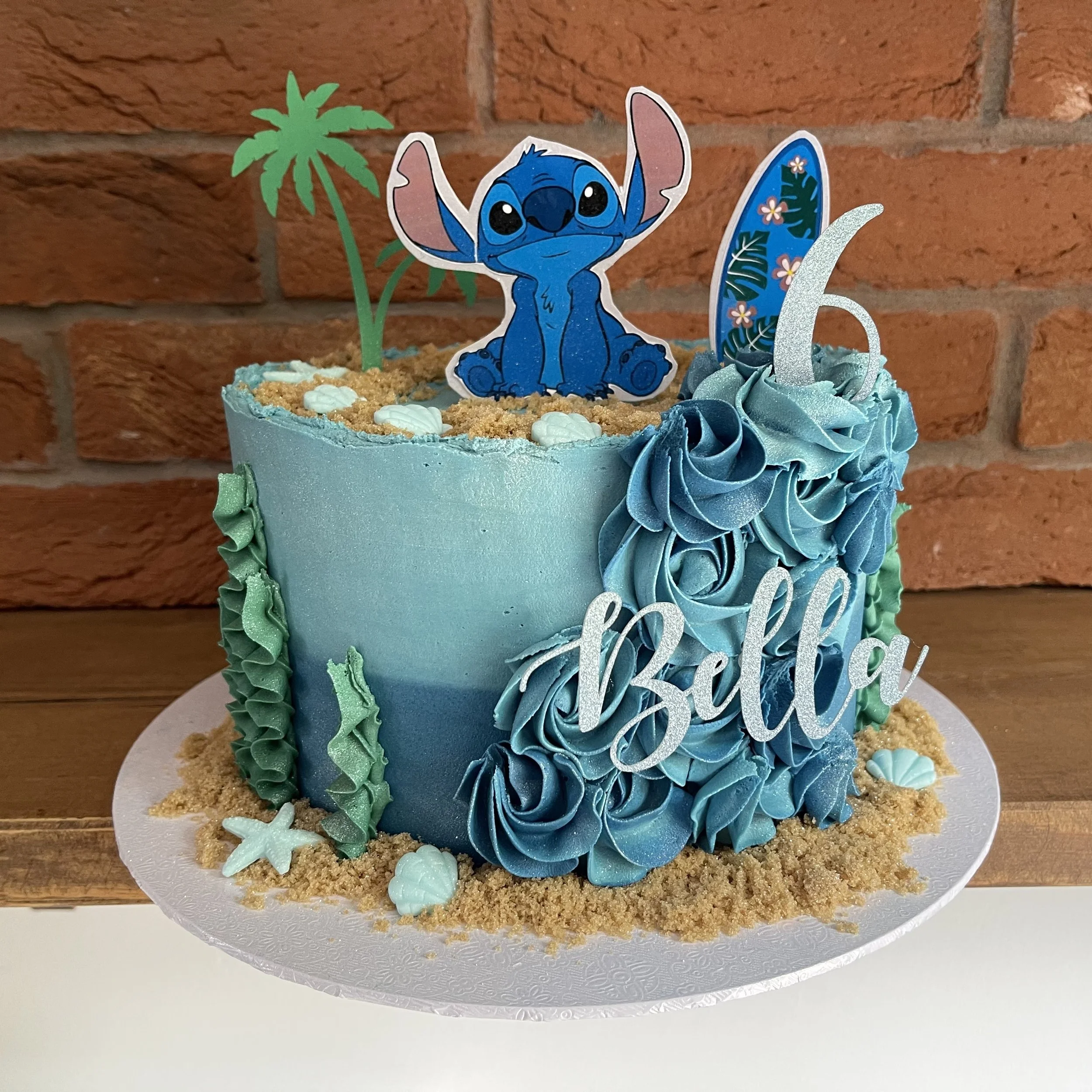 A blue ocean-themed birthday cake with a "Lilo & Stitch" character topper, palm tree decoration, sea-themed frosting, and "Bella" written on it. A "6" candle is also present.