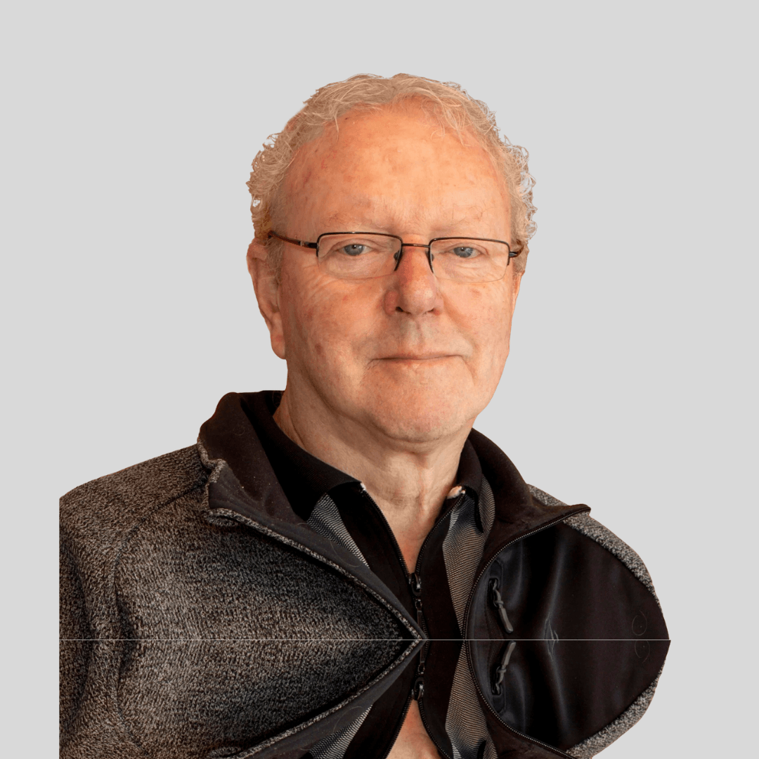 Portrait of an older man with glasses, wearing a black and gray jacket, posing against a plain light gray background.