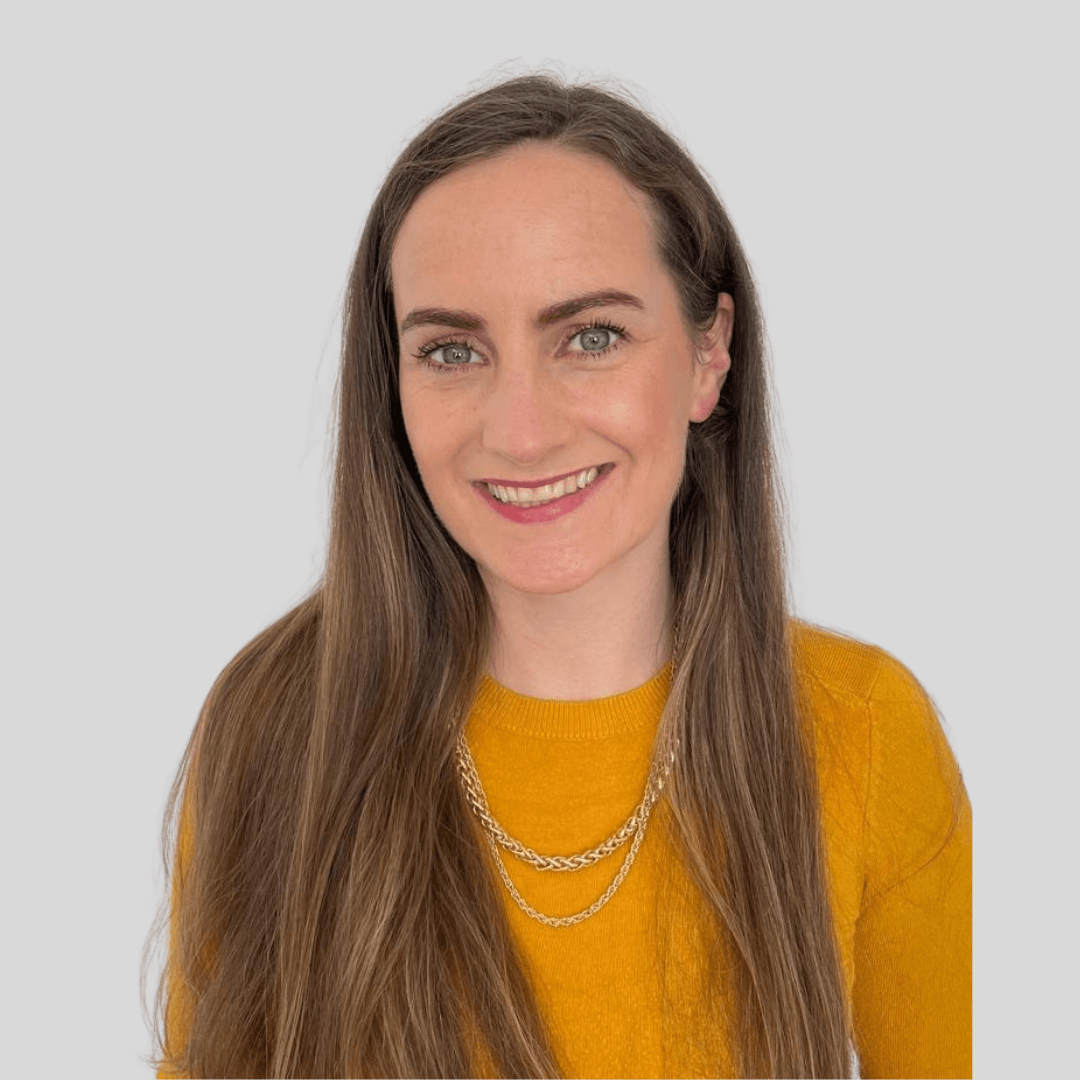 Portrait of a woman with long brown hair, blue eyes, and fair skin, smiling, wearing a mustard yellow sweater and layered gold necklaces against a light gray background.