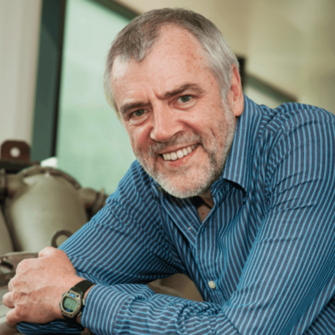 Senior man with gray hair and beard smiling, wearing a blue striped shirt, sitting indoors with a window in the background.