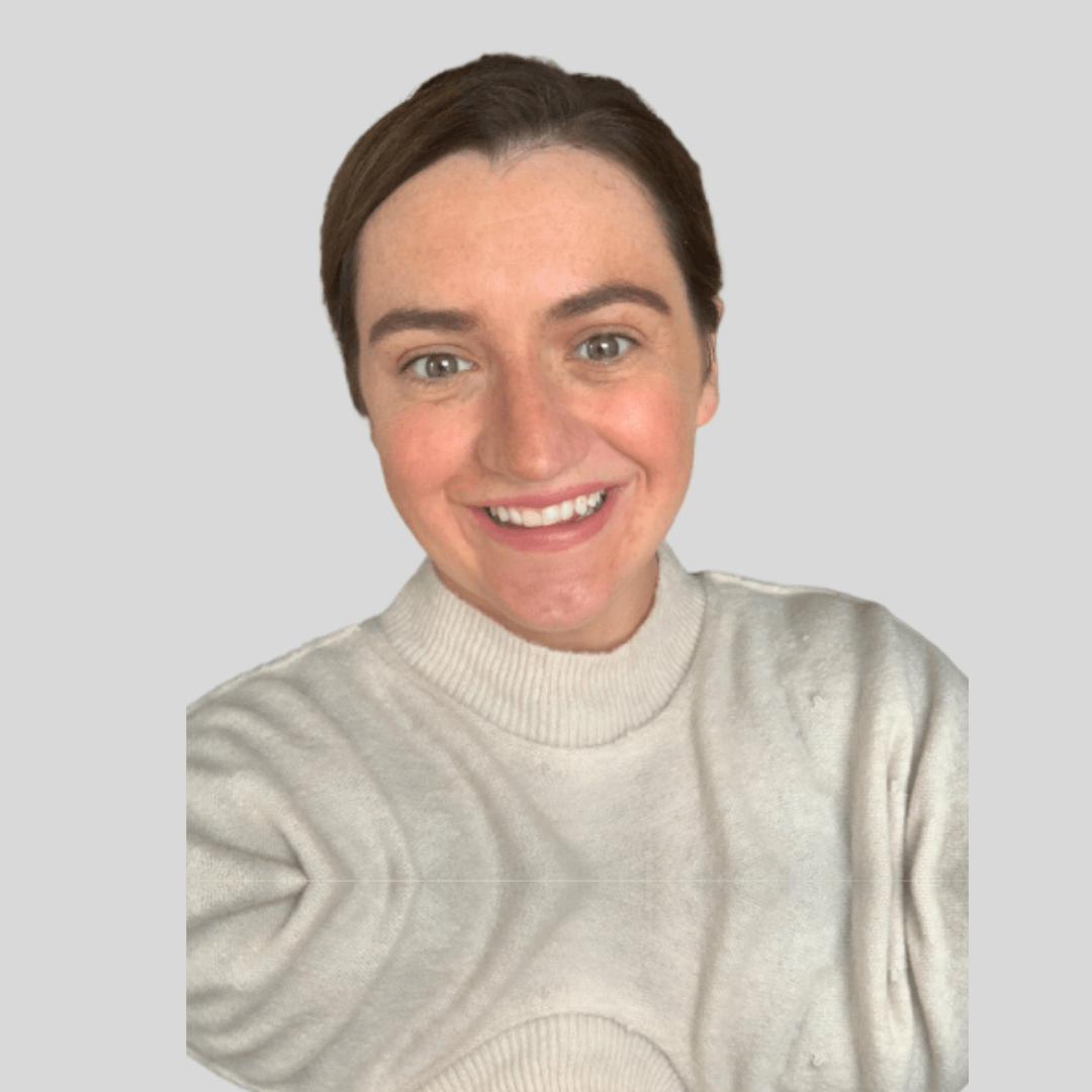 Smiling woman with short brown hair wearing a beige sweater, taking a selfie against a plain light gray background.
