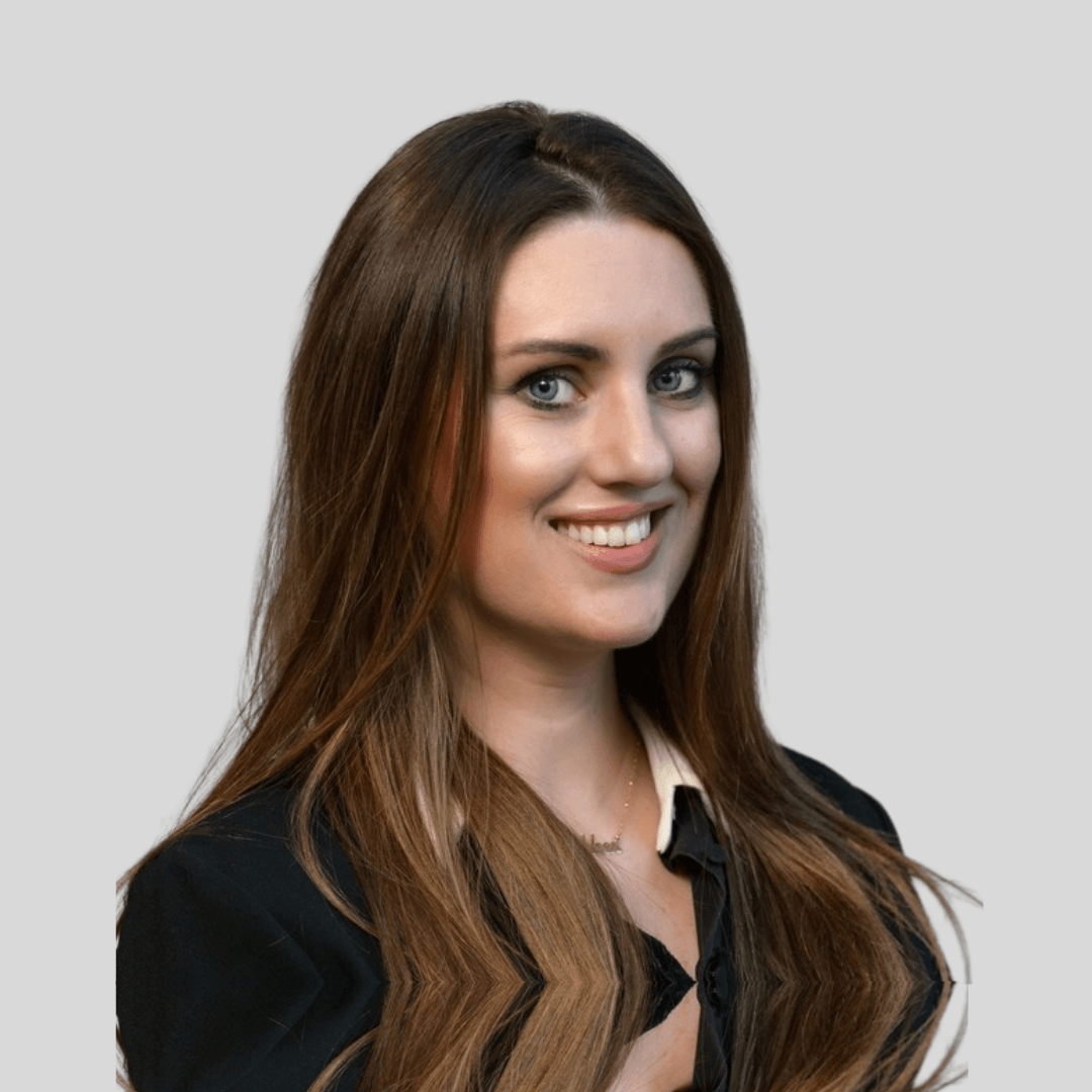 Portrait of a smiling woman with long brown hair, blue eyes, and fair skin, wearing a black top and a necklace, against a plain light gray background.