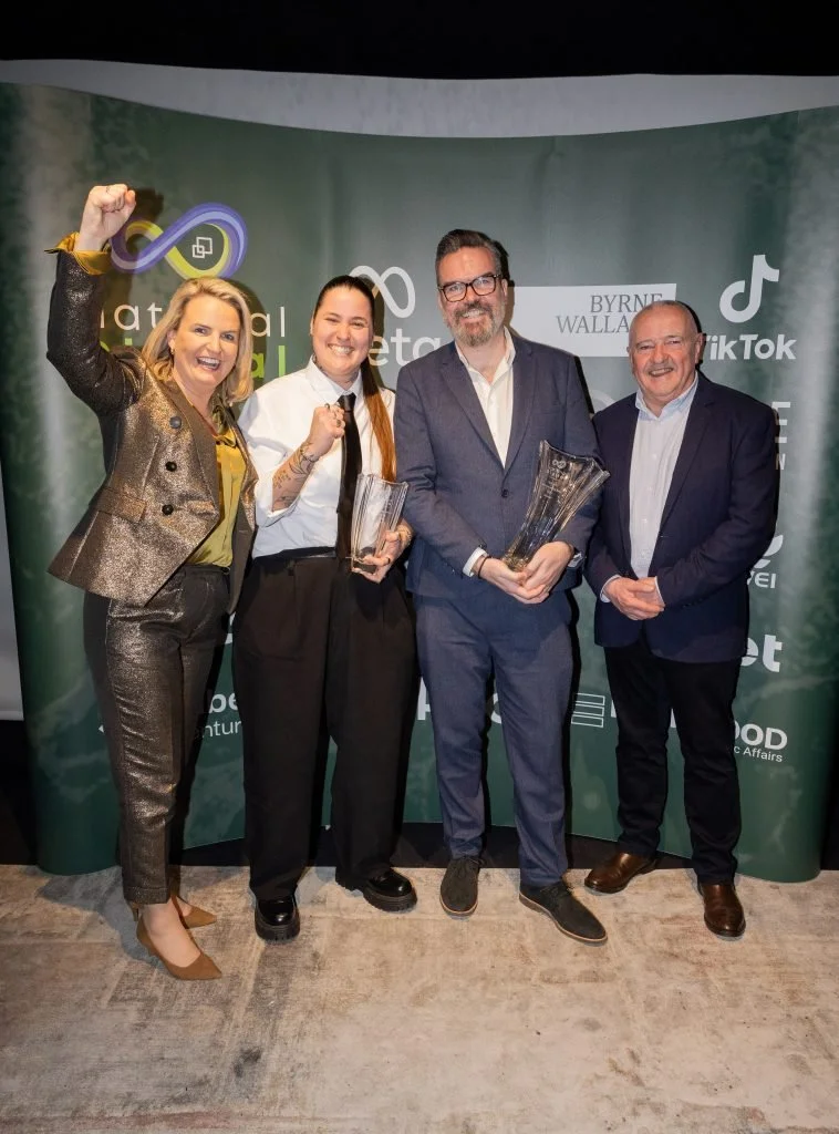 Four people standing together at an award ceremony, holding trophies and celebrating, with a green backdrop featuring various logos and names.