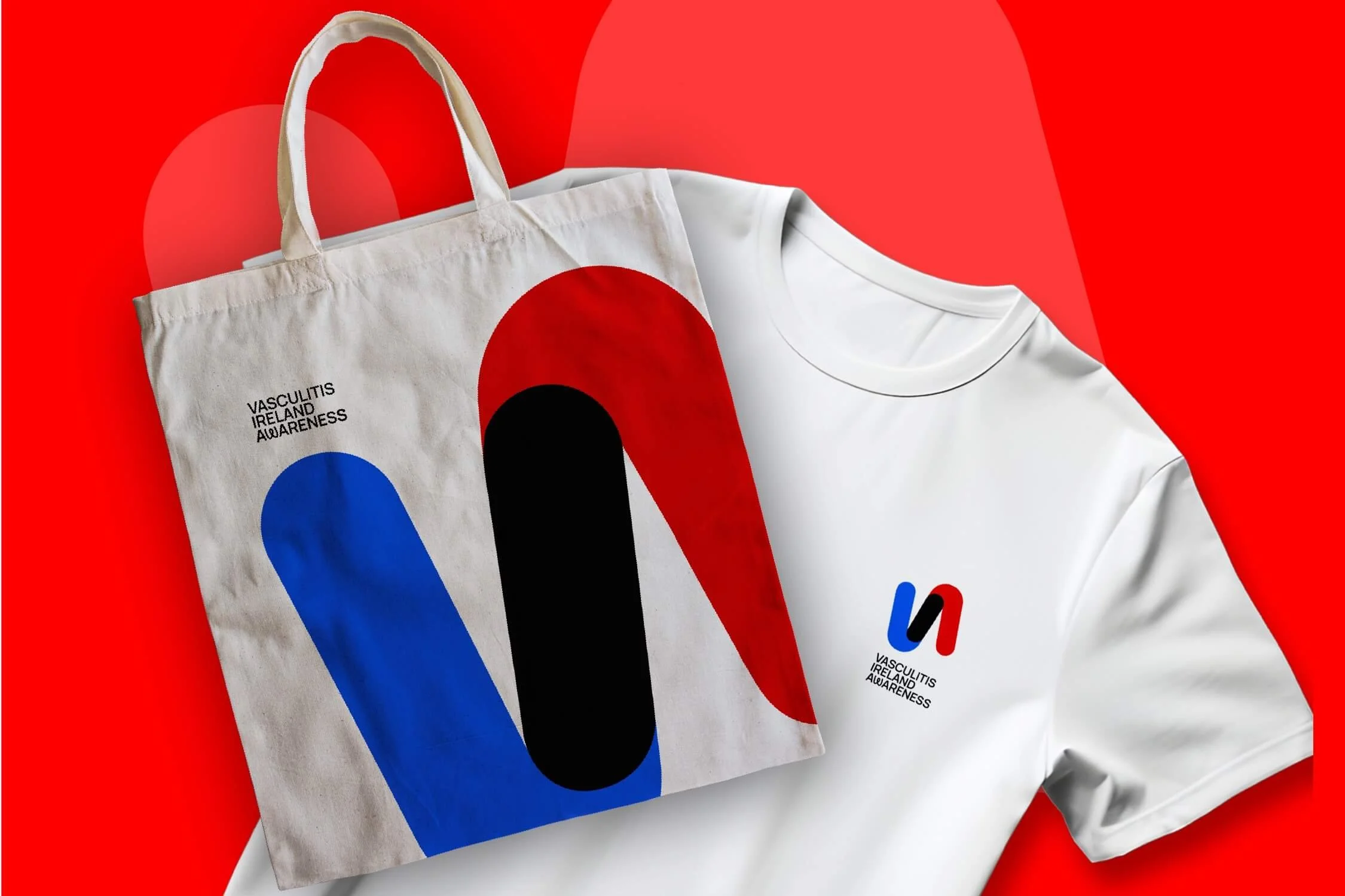 A white tote bag and a white T-shirt with Vascultis Ireland Awareness logos placed on a red background. The tote bag features large abstract shapes in red, black, and blue, with text in black.