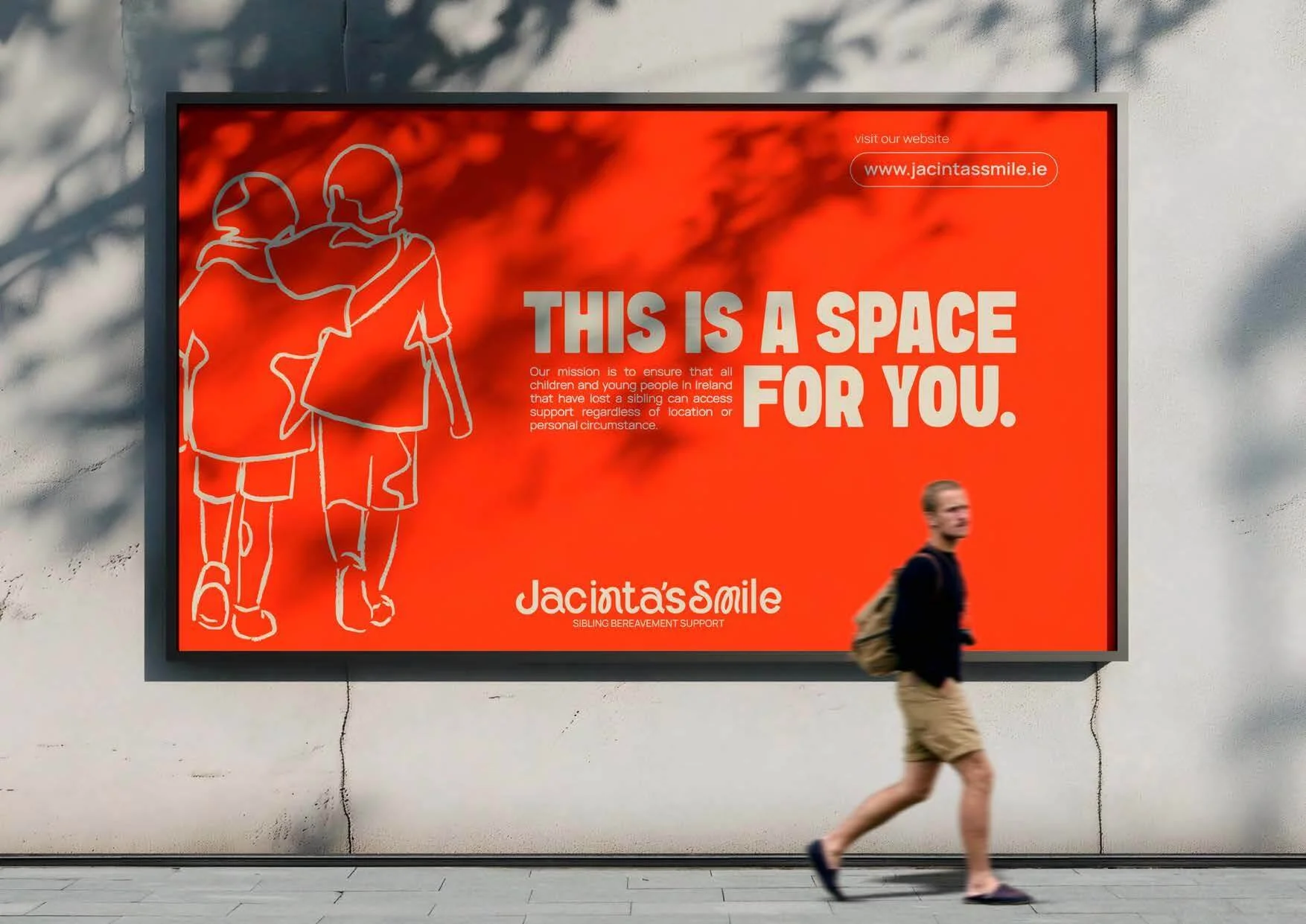 A billboard with an orange background shows white line art of two children walking with arms around each other. Large text reads, 'THIS IS A SPACE FOR YOU.' Additional smaller text explains the mission of Jacinta's Smile, a sibling bereavement support organization. A woman walks in front of the billboard along the sidewalk.