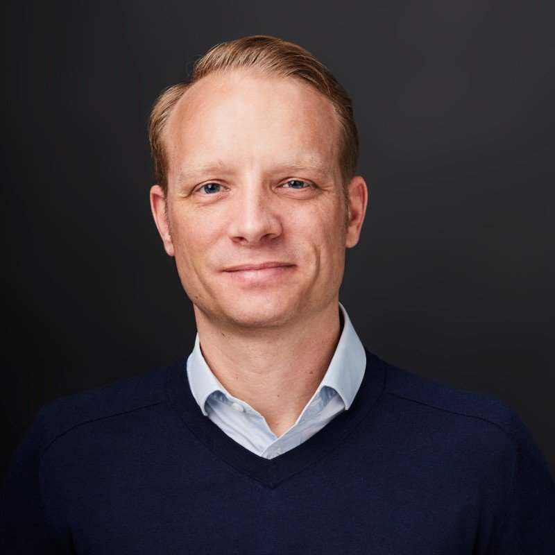 Porträt eines Mannes, Jens Erasmus, mit blondem Haar in Business-Kleidung vor dunklem Hintergrund.