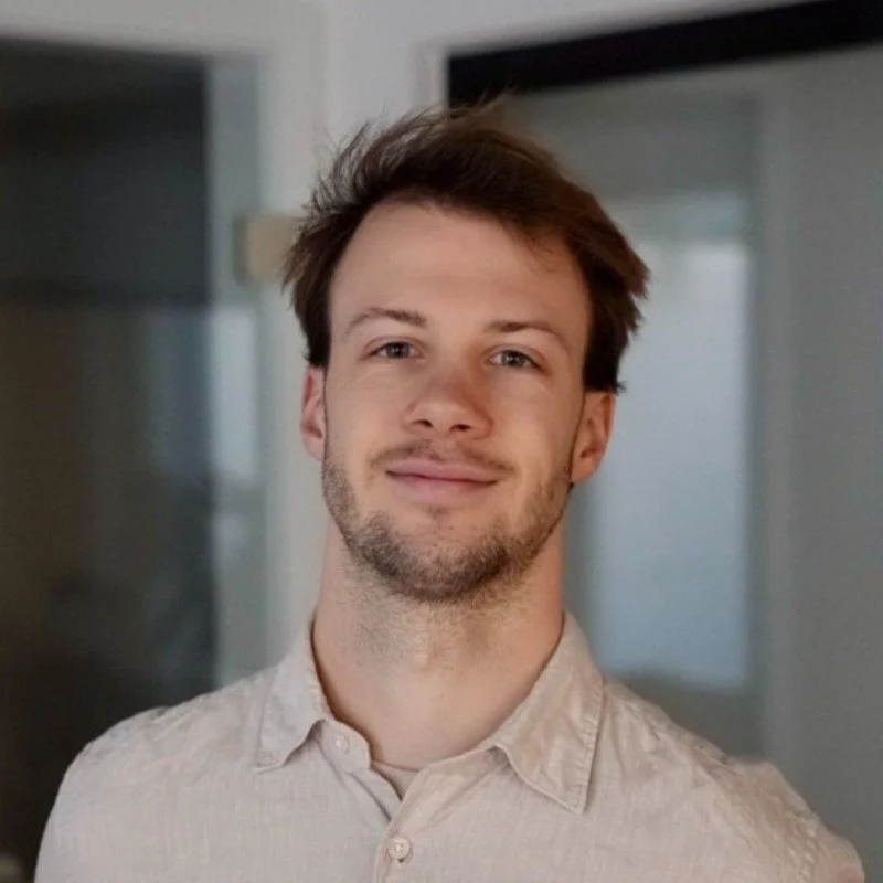 Ein junger Mann mit braunem Haar und Bart, trägt ein helles Hemd, lächelt leicht, im Innenraum.