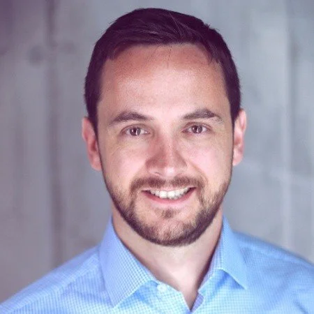 Porträt von Willi Tscheschner, eines lächelnden Mannes mit dunklem Haar, Bart und blauer Hemd vor einem grauen Hintergrund.