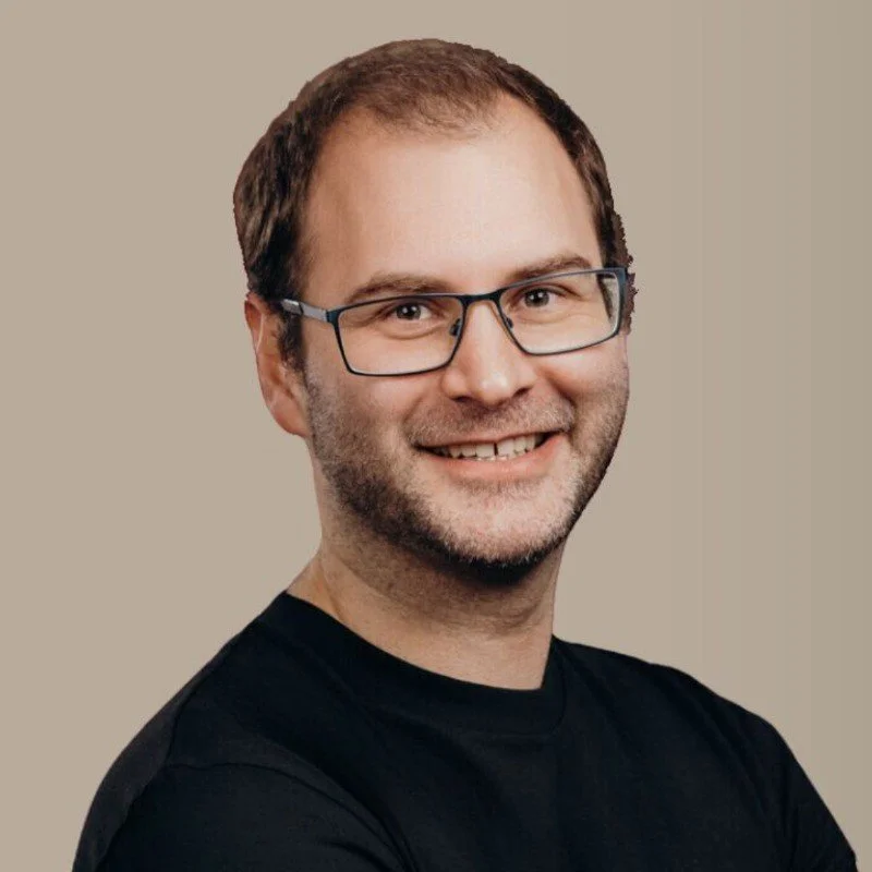 Ein Mann, Torben Schreiter, mit Brille, dunklem Haar und Bart, trägt ein schwarzes T-Shirt, vor neutralem Hintergrund, lächelnd.