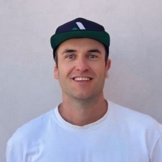 Ein Mann, Julius Göllner, mit dunkelblau-grünem Cap und weißem T-Shirt, lächelnd vor hellem Hintergrund.