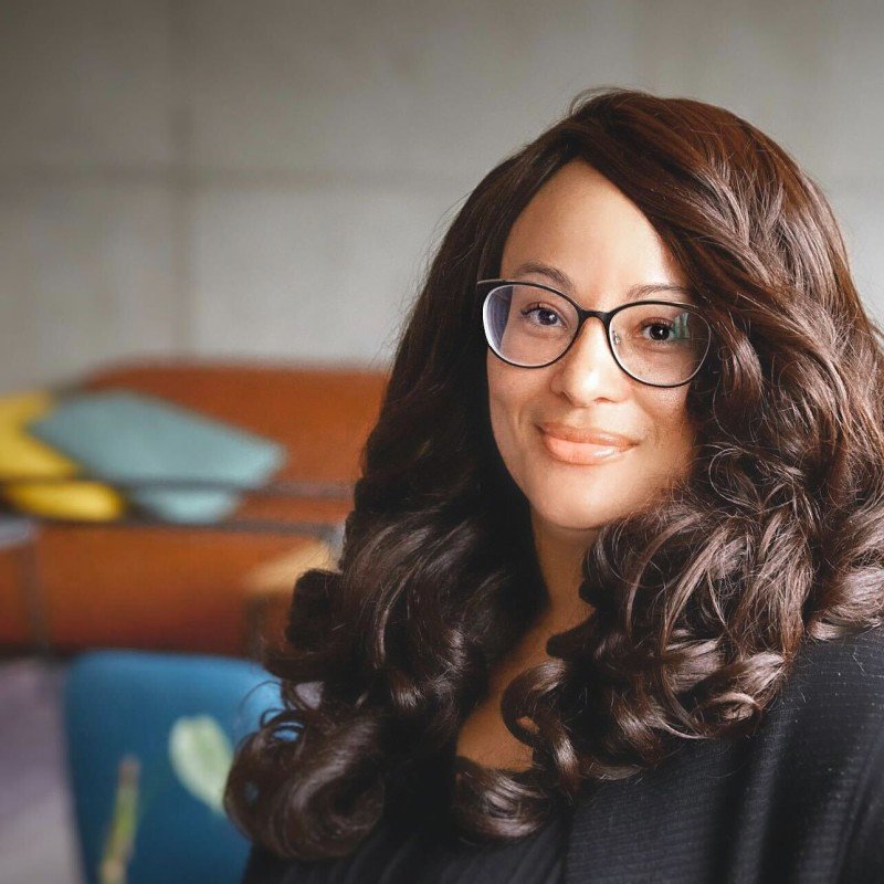 Eine Frau, Angeley Mullins, mit langen, lockigen braunen Haaren, trägt eine Brille und lächelt freundlich.