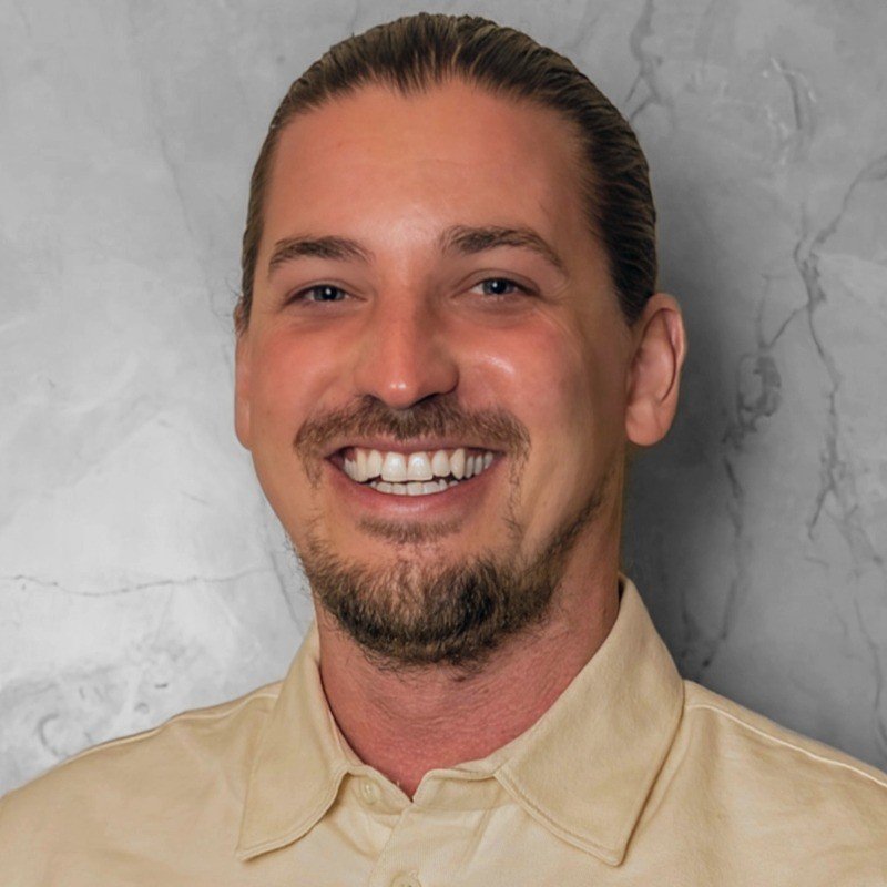 Ein lächelnder Mann, Nicolas Peters, mit braunem Haar, kurzer Bart und goatee, trägt ein hellbeiges Hemd, vor einer grauen Marmorwand.