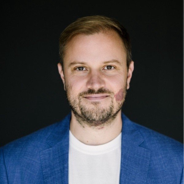 Porträt eines Mannes, Gero Decker, mit kurzen braunen Haaren, Bart, trägt eine blaue Jacke und ein weißes Hemd, steht vor dunklem Hintergrund.