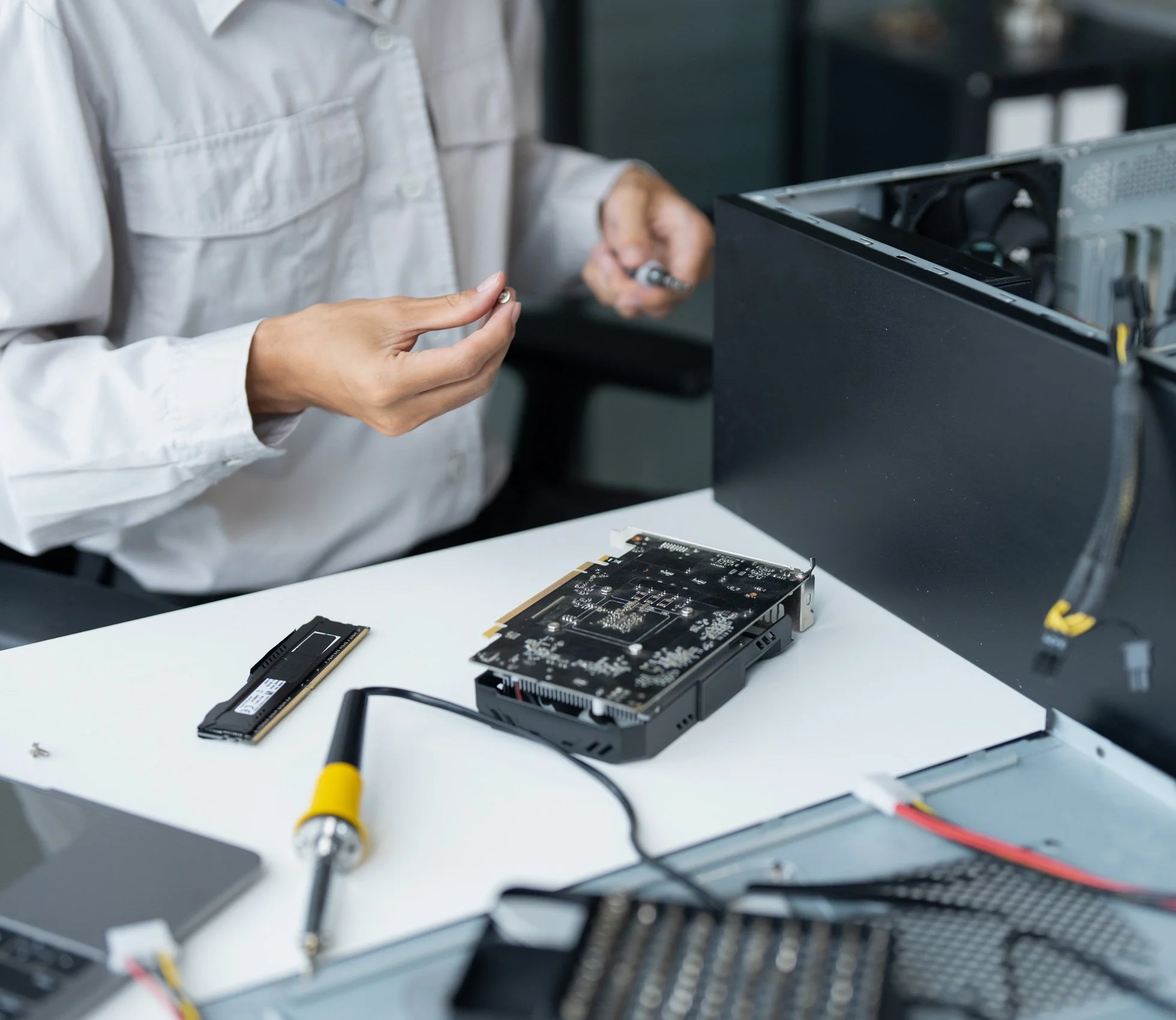 Person working on computer hardware, handling a small circuit with tools, with various electronic components on a white desk.