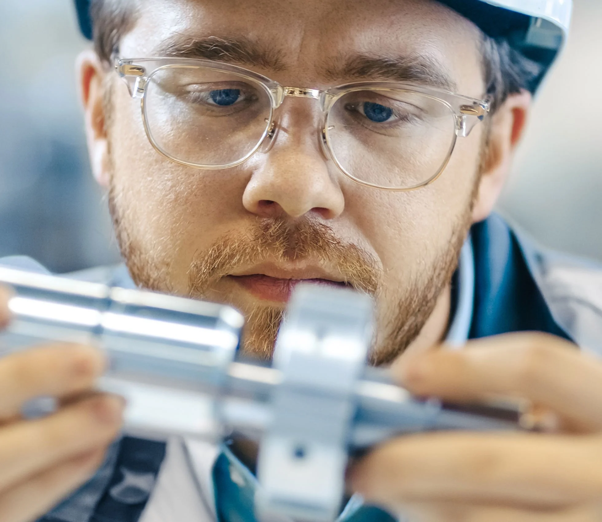 A man wearing safety glasses and a helmet works with a metallic object, focusing intently on his task.