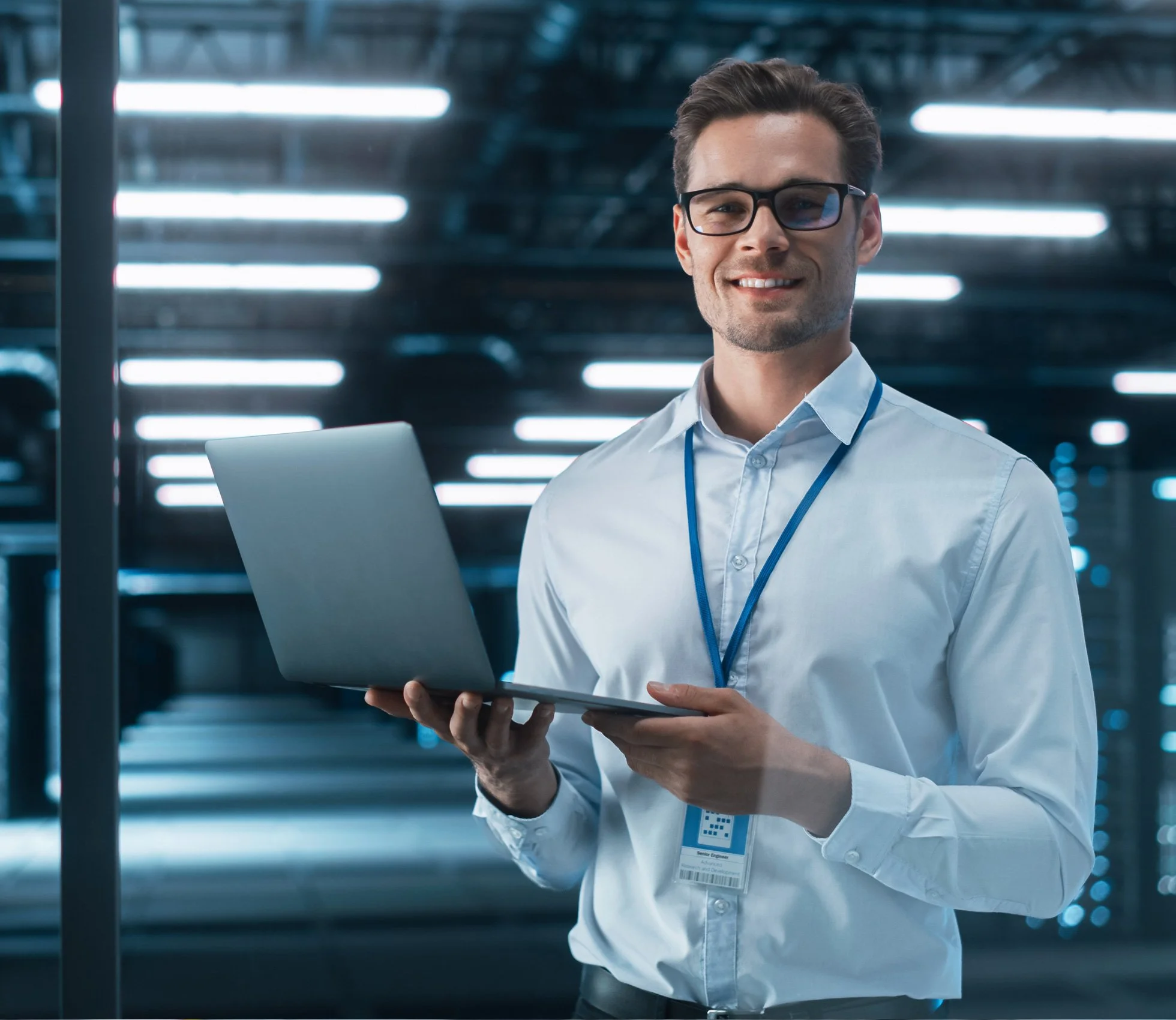 A man wearing glasses, a white shirt, and a blue lanyard, holding a laptop in a data center with server racks and bright overhead lights.