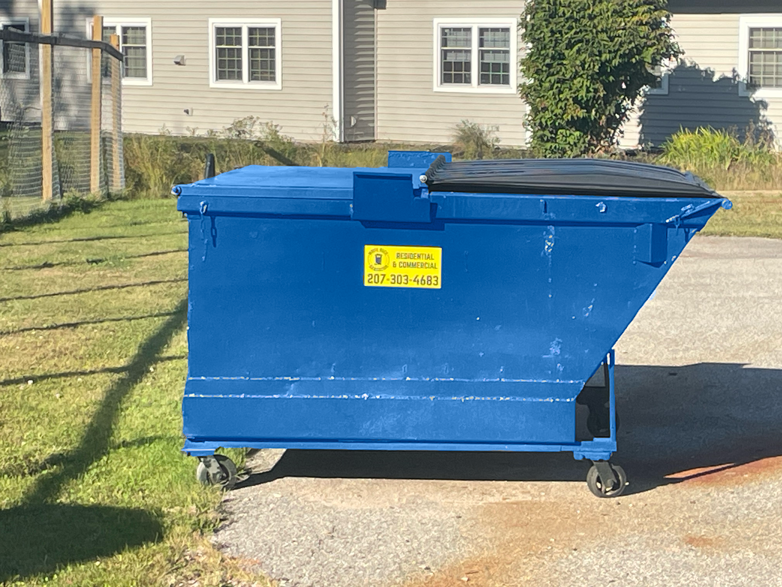 Blue large residential and commercial trash dumpster with a yellow sticker outdoors with grass, houses, trees, and a fence in the background in Gorham, ME. Commercial garbage service residential trash service