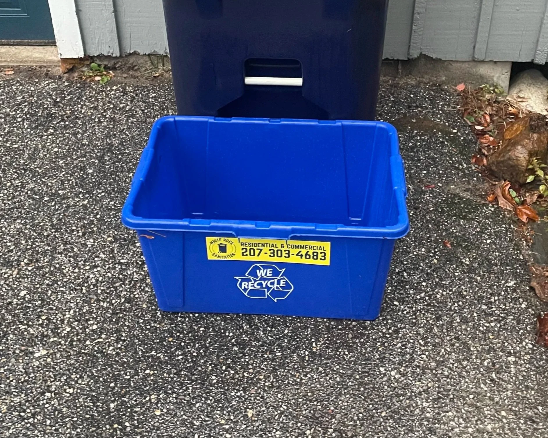 Blue recycling bin placed outside on a gravel ground near a building, with a black lid and a yellow sticker displaying a phone number and information about residential and commercial recycling services for White Rock Sanitation in Gorham, Maine.