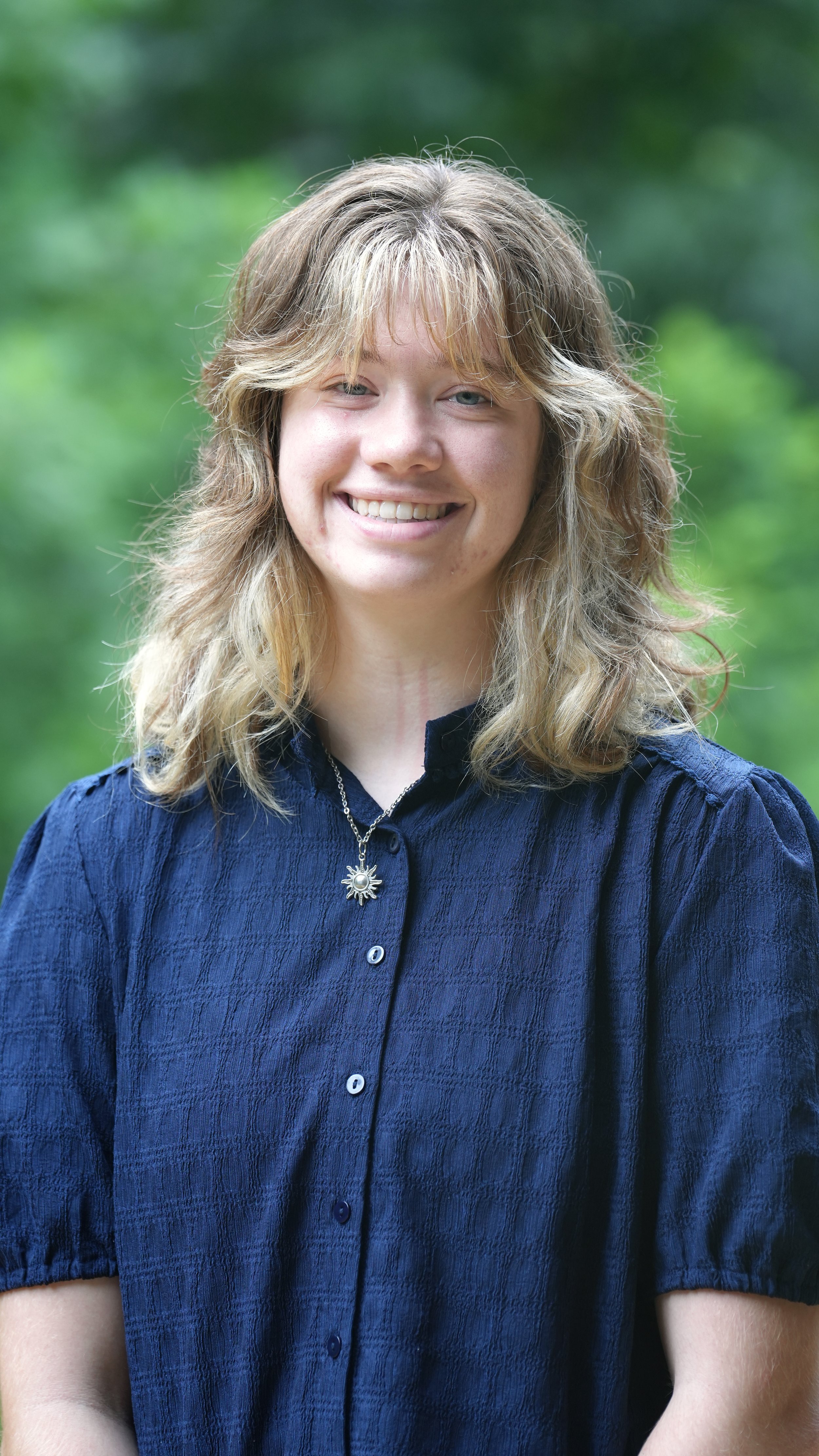Close-up of a woman with reddish-brown hair wearing a dark gray jacket, smiling, with a background of green trees.