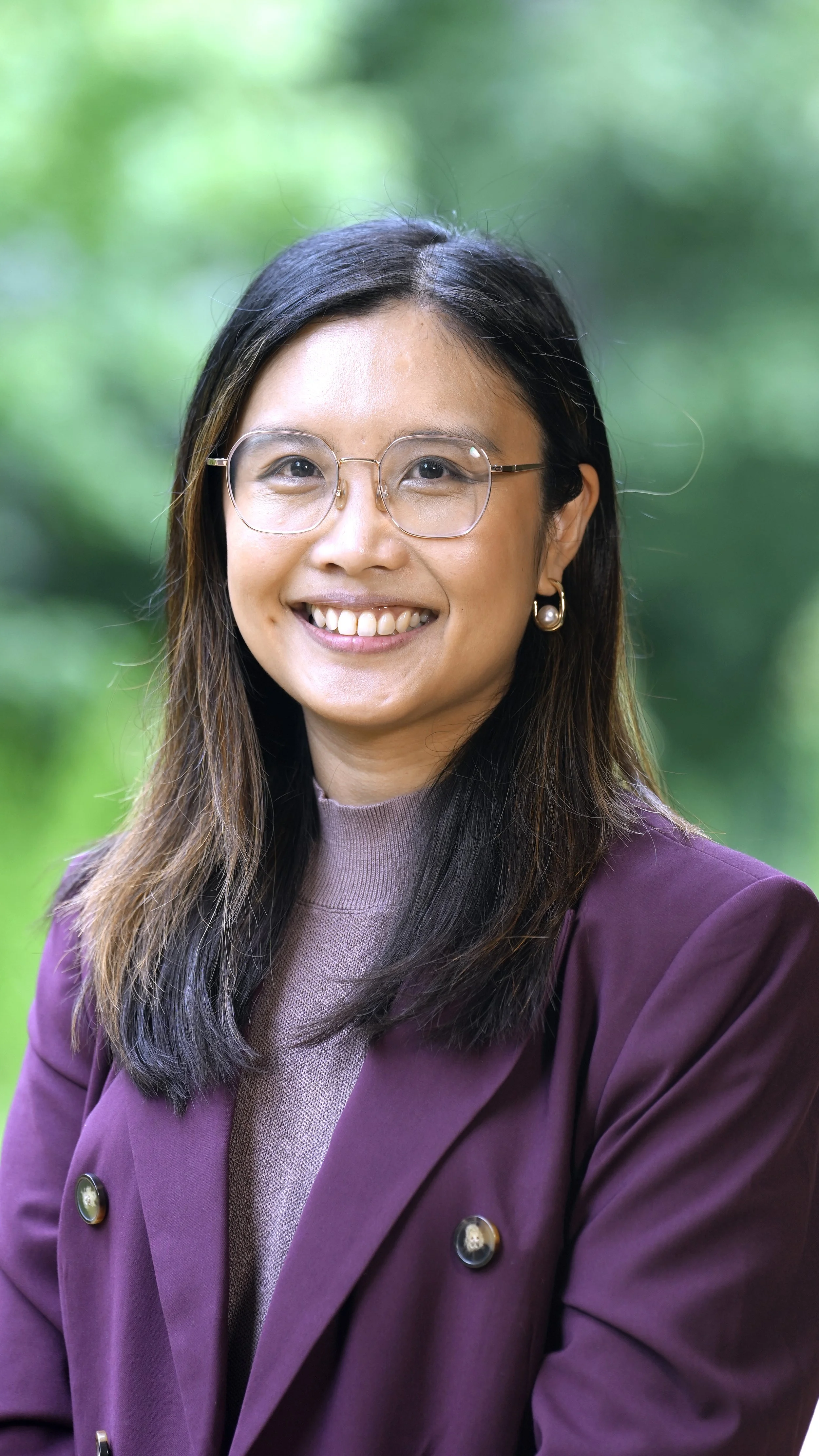 Portrait of a woman with glasses smiling outdoors with a blurred green background.