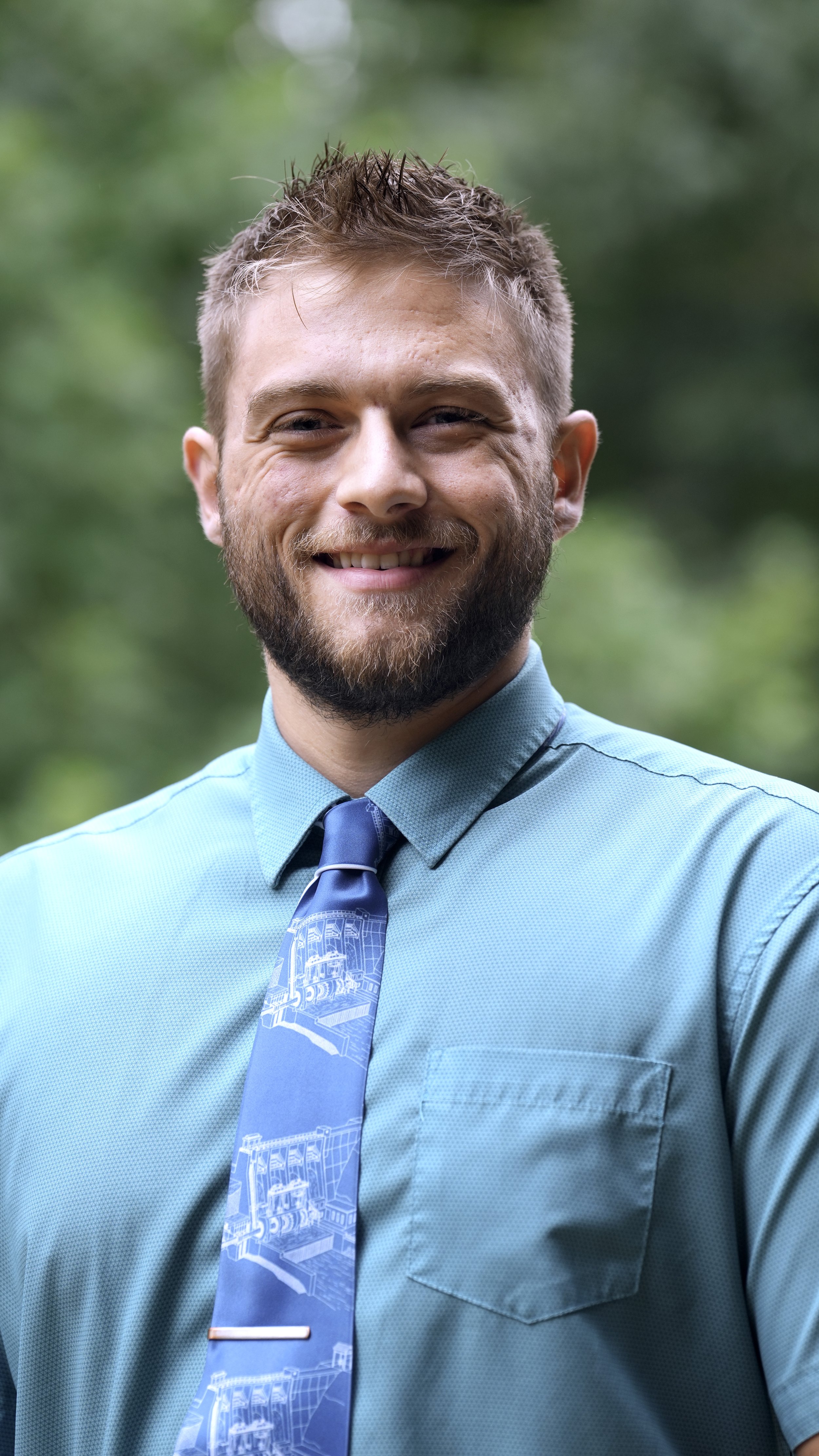 A young man with light skin, curly hair, and a beard smiling outdoors with a blurred green leafy background. He is wearing a dark blue shirt with a small repeating pattern and a light yellow tie with dinosaur illustrations.