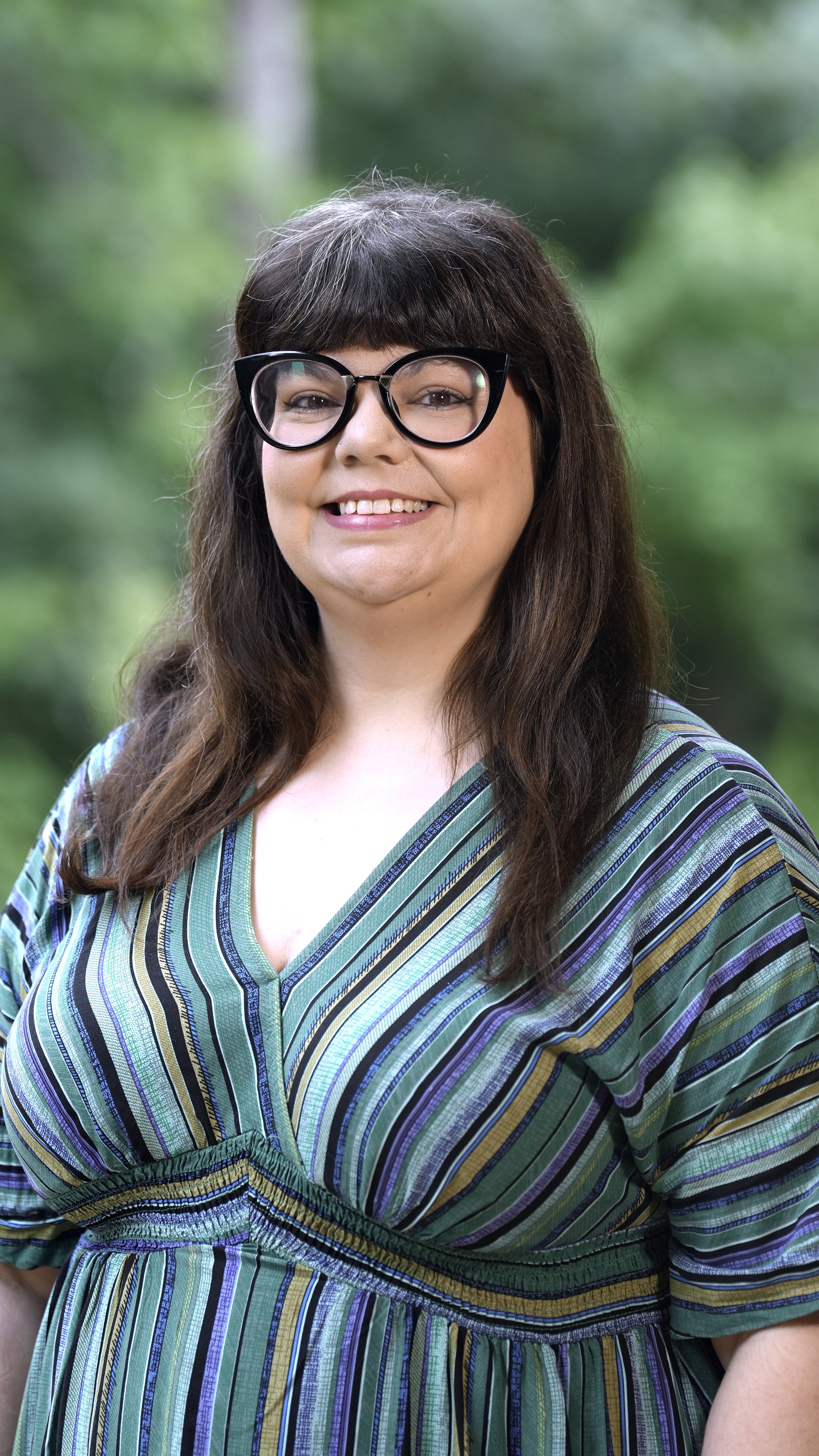 A smiling woman with brown hair and wearing large black glasses, a green top, a denim jacket, and a silver necklace with a pendant, standing outdoors with green foliage in the background.