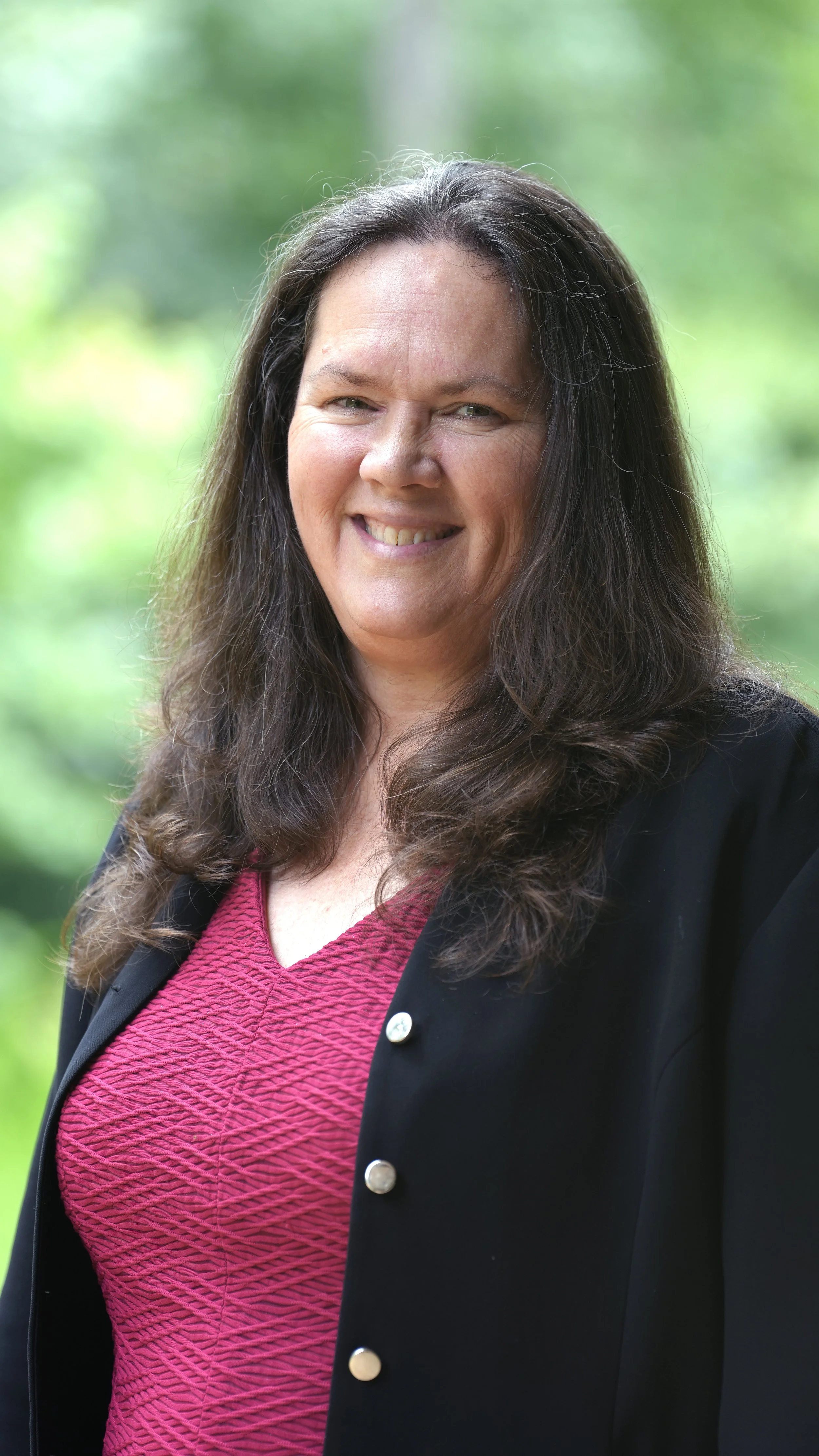 A woman with short brown hair, smiling, wearing a pink polka-dot blouse and black blazer, outdoors with blurred green trees in the background.