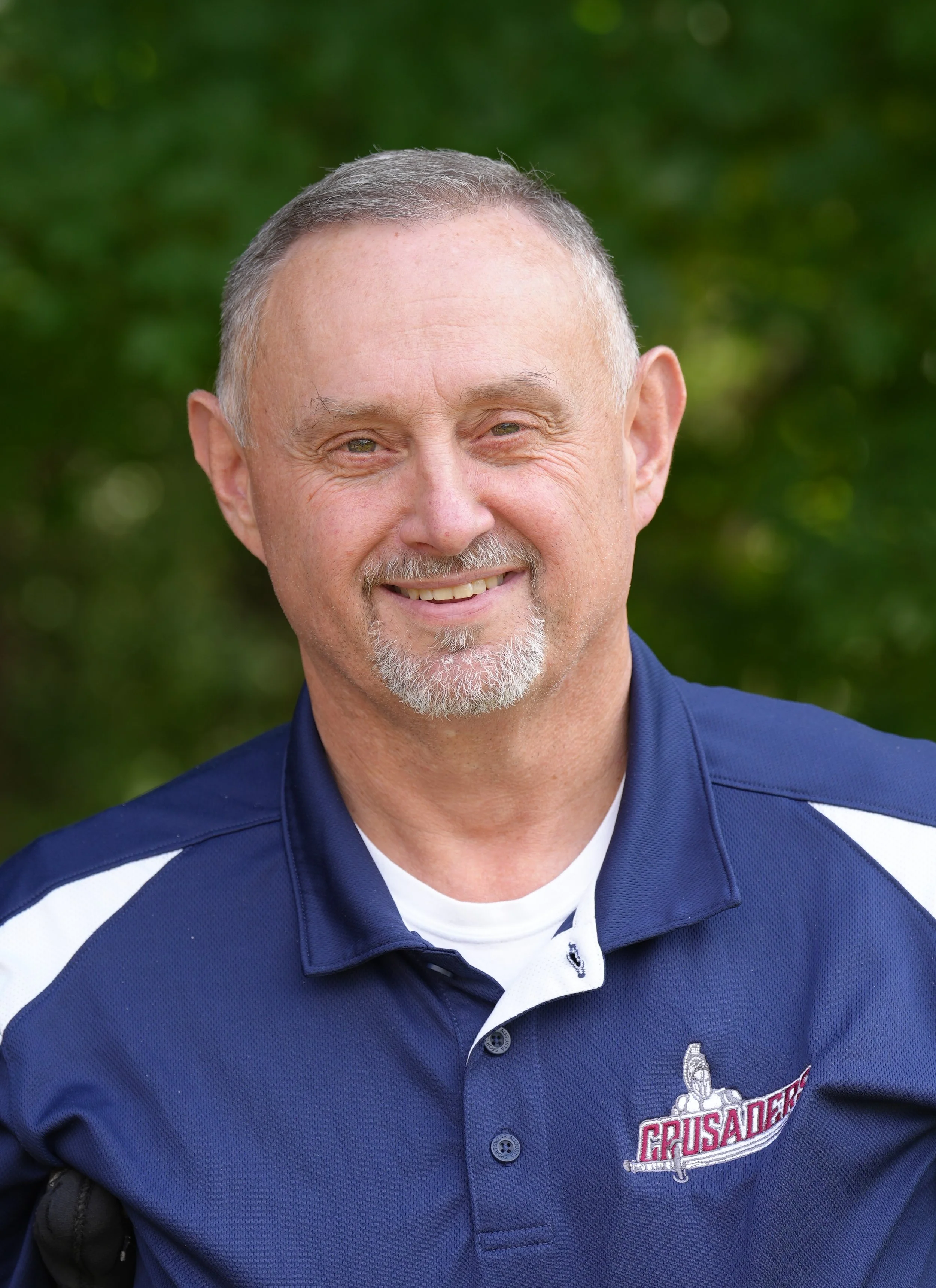 A middle-aged man with short, gray hair and a beard, wearing a maroon polo shirt with the logo of a bulldog and the word 'Crusaders' on it. He is outdoors with blurred green foliage in the background.