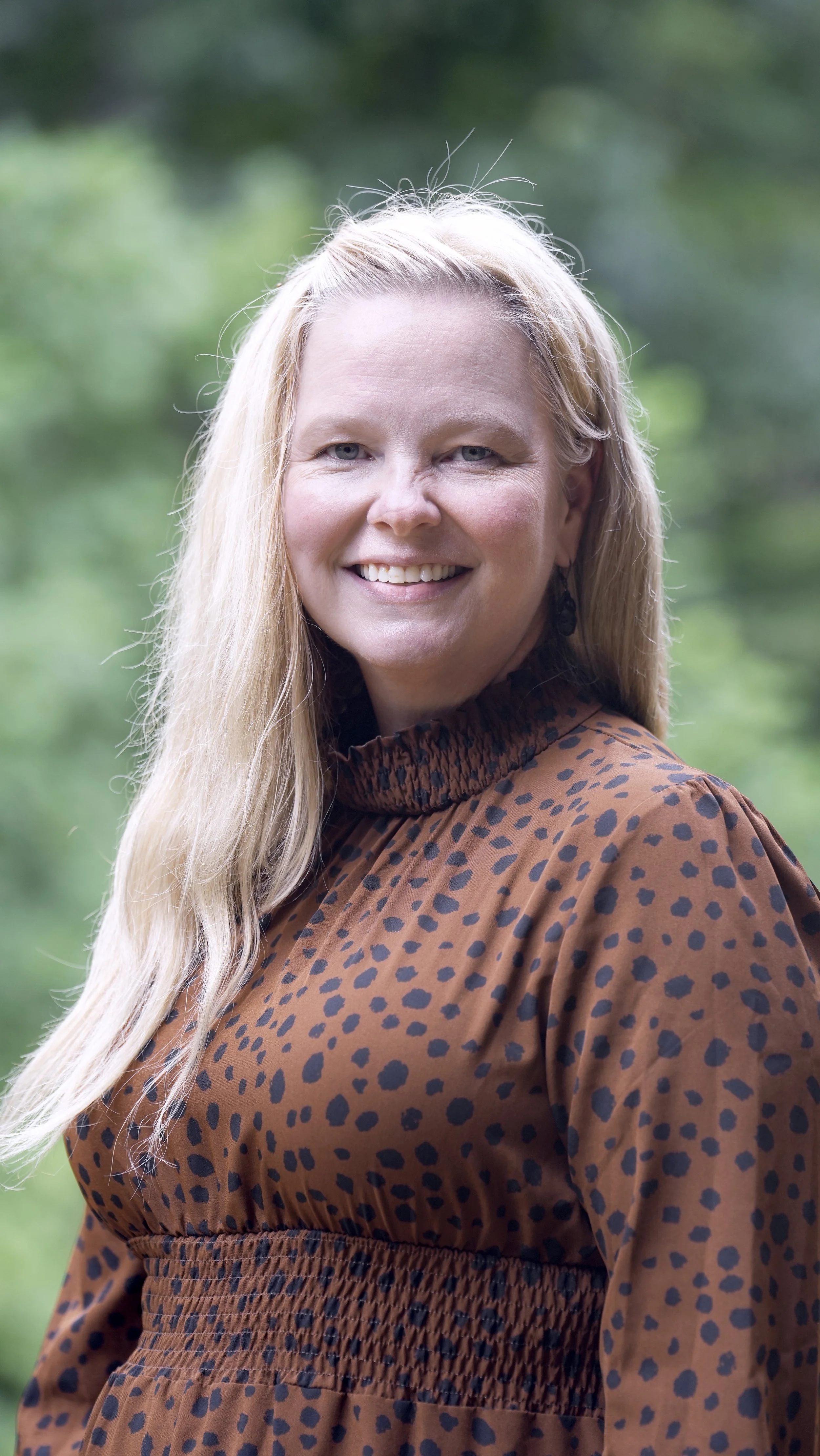 Close-up of a smiling woman with long curly blonde hair, wearing yellow earrings and a mustard yellow cardigan, outdoors with blurred green foliage in the background.