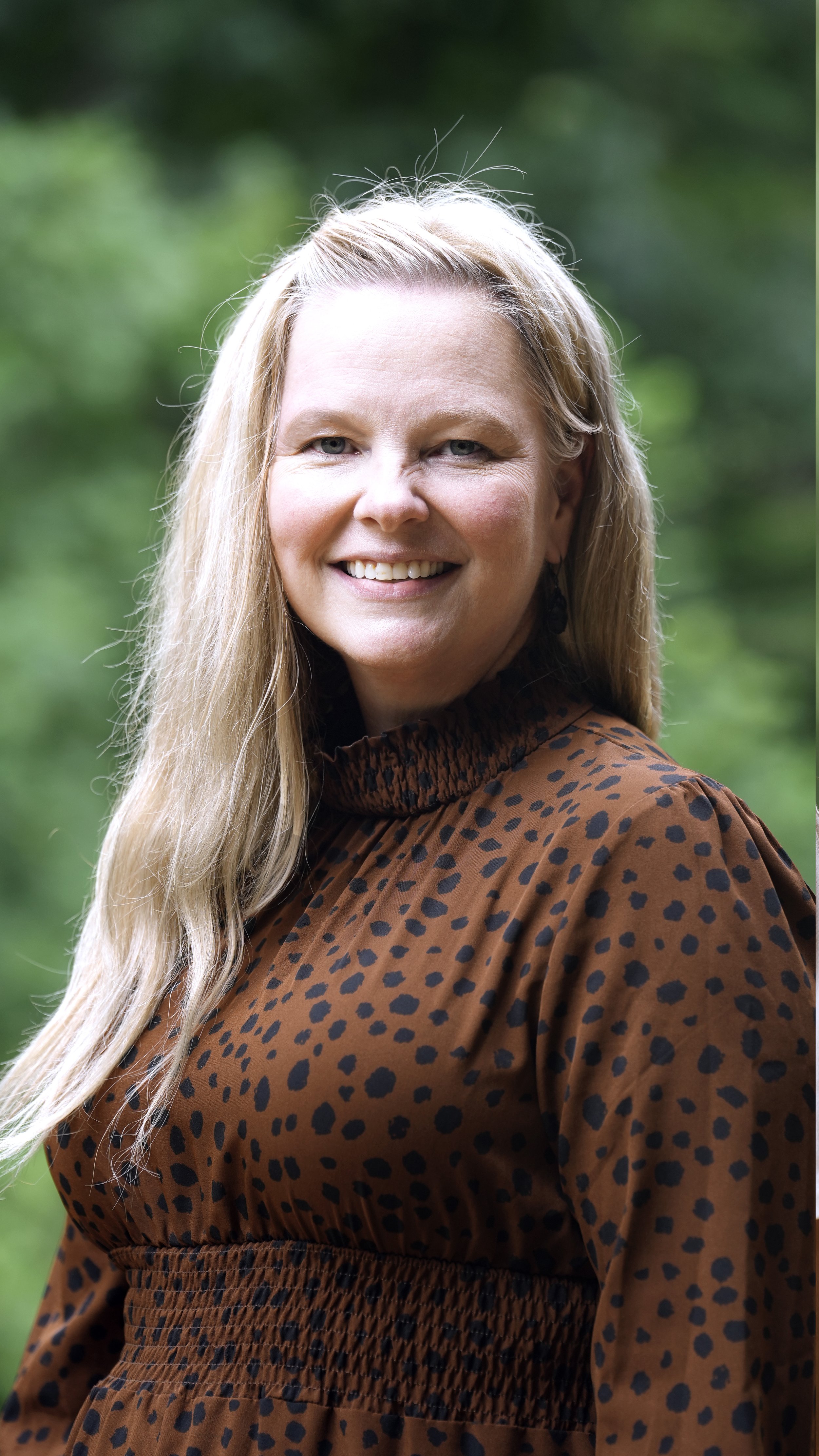 Close-up of a smiling woman with long curly blonde hair, wearing yellow earrings and a mustard yellow cardigan, outdoors with blurred green foliage in the background.