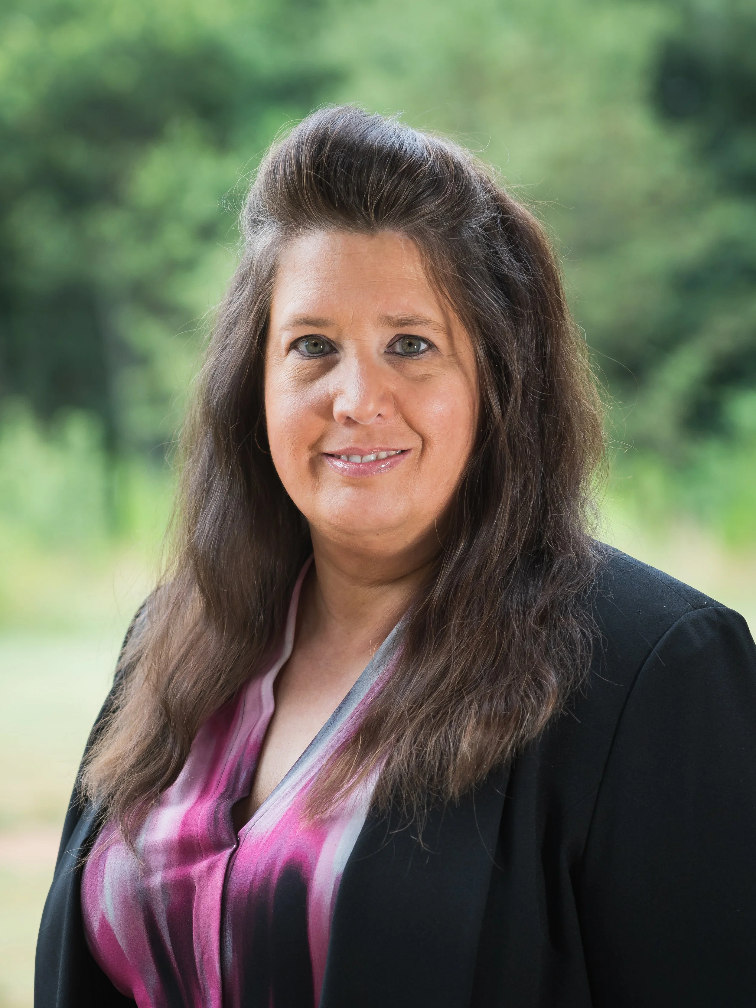 Close-up portrait of a woman with long brown hair, wearing a black blazer over a pink and purple blouse, standing outdoors with blurred green trees in the background.