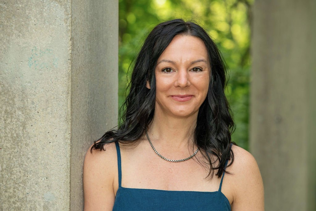 A woman with shoulder-length black hair, wearing a blue sleeveless top and a necklace, standing outdoors near concrete pillars with green trees in the background.