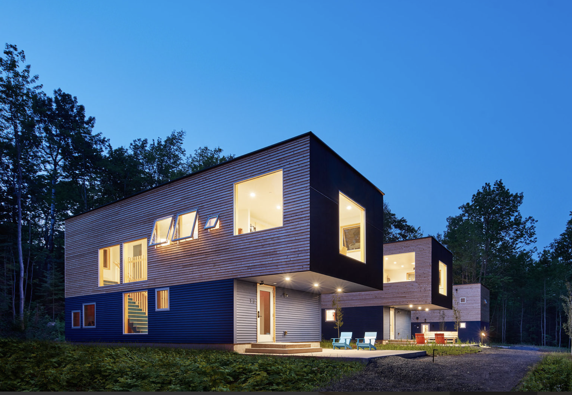 Modern multi-story house with large windows illuminated at night, set in a wooded area, with outdoor seating on a patio.