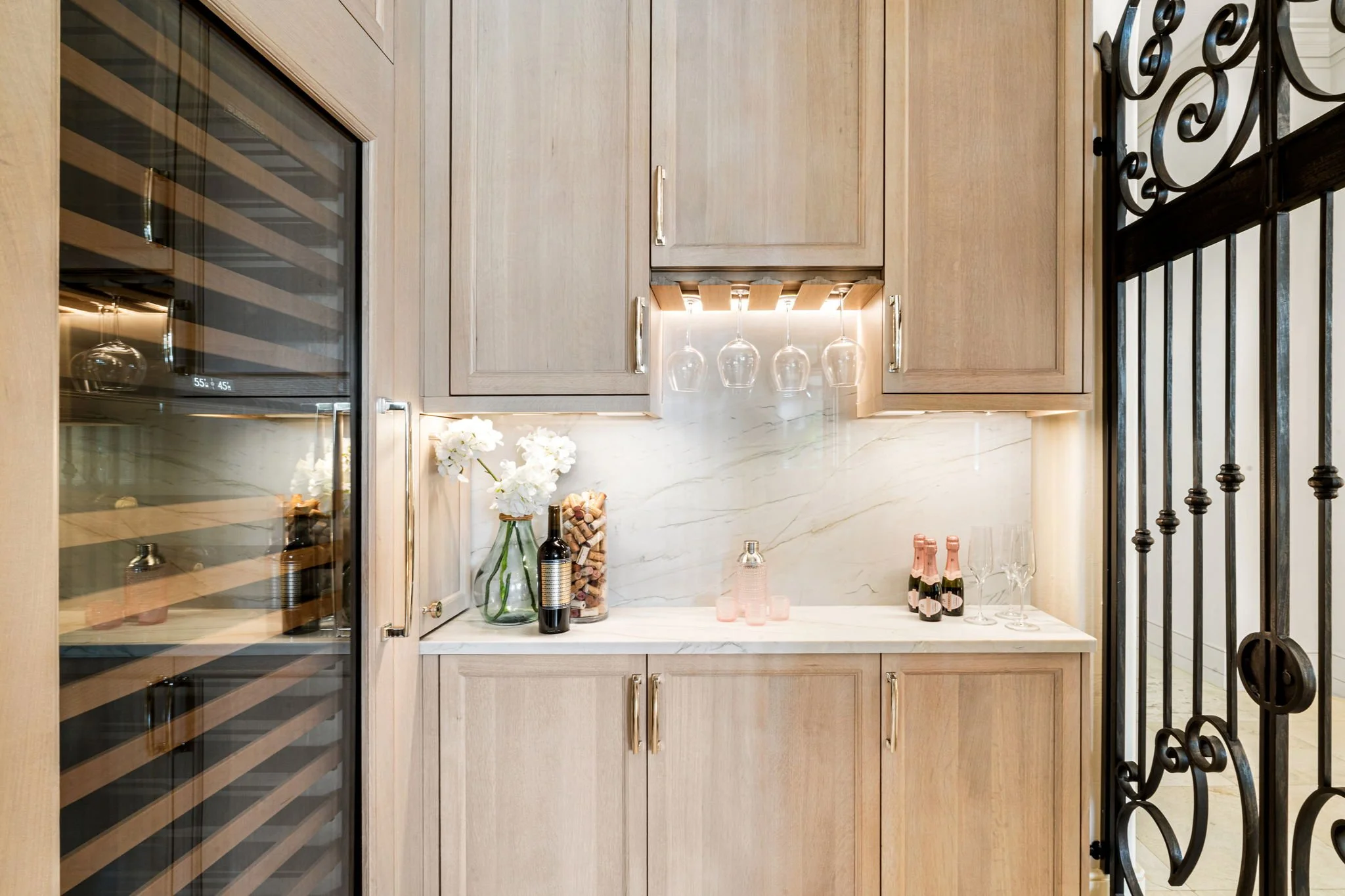 A built-in wine cooler with glass door on the left, wooden cabinets with brass handles, a white marble countertop with a vase of white flowers, a bottle of wine, and five wine glasses hanging upside down under the cabinet. There are champagne bottles