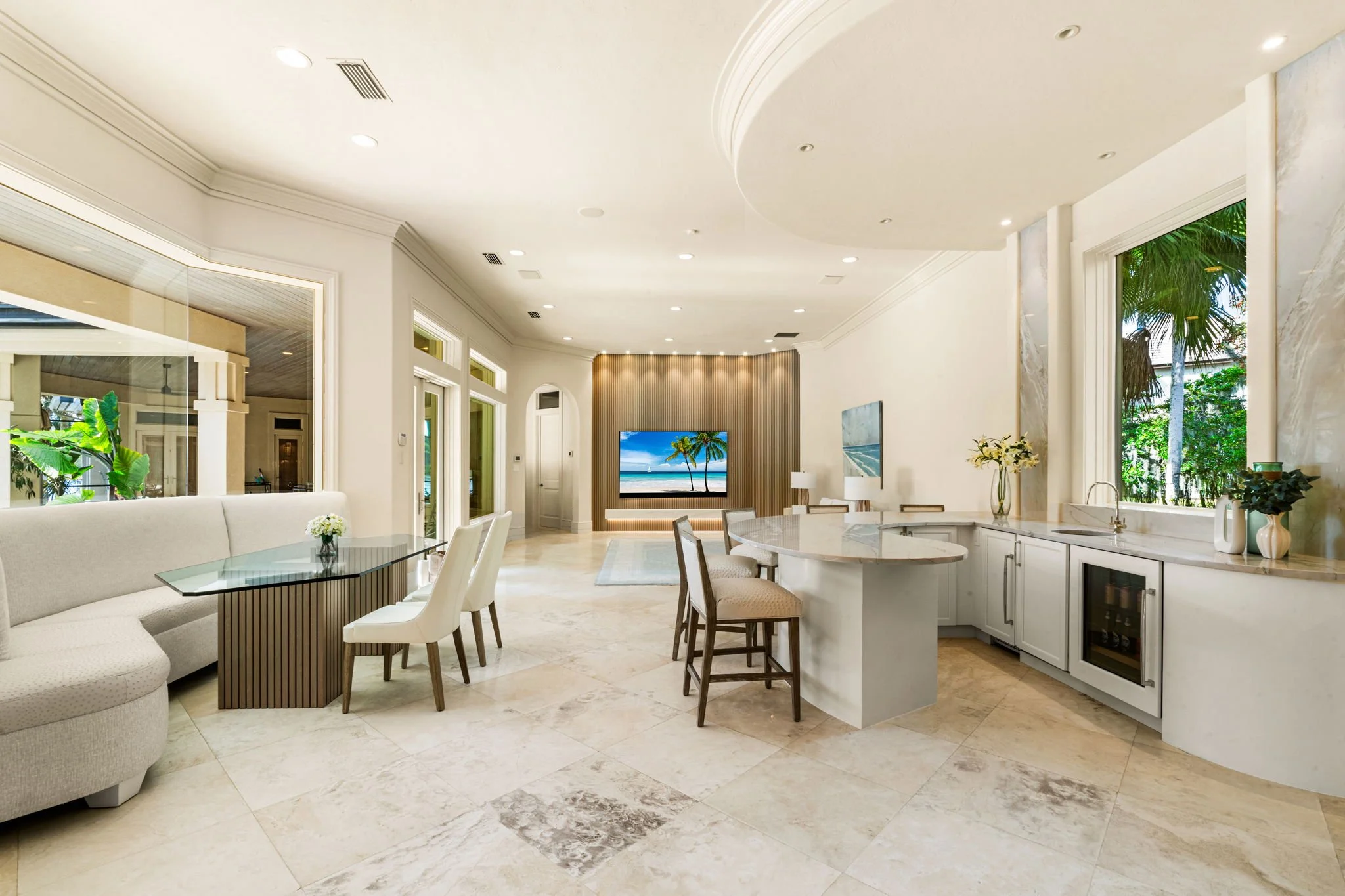 Spacious, modern living area with a kitchen, dining space, and a sitting area on tile flooring. Large windows with views of tropical trees, a flat-screen TV, and contemporary furnishings.