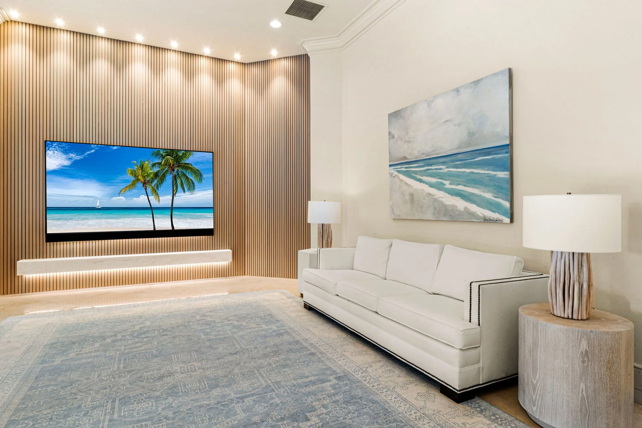 Modern living room with white sofa, wooden side table, and lamps, with a painting of a beach scene on the wall, and a large TV displaying a tropical beach with palm trees.