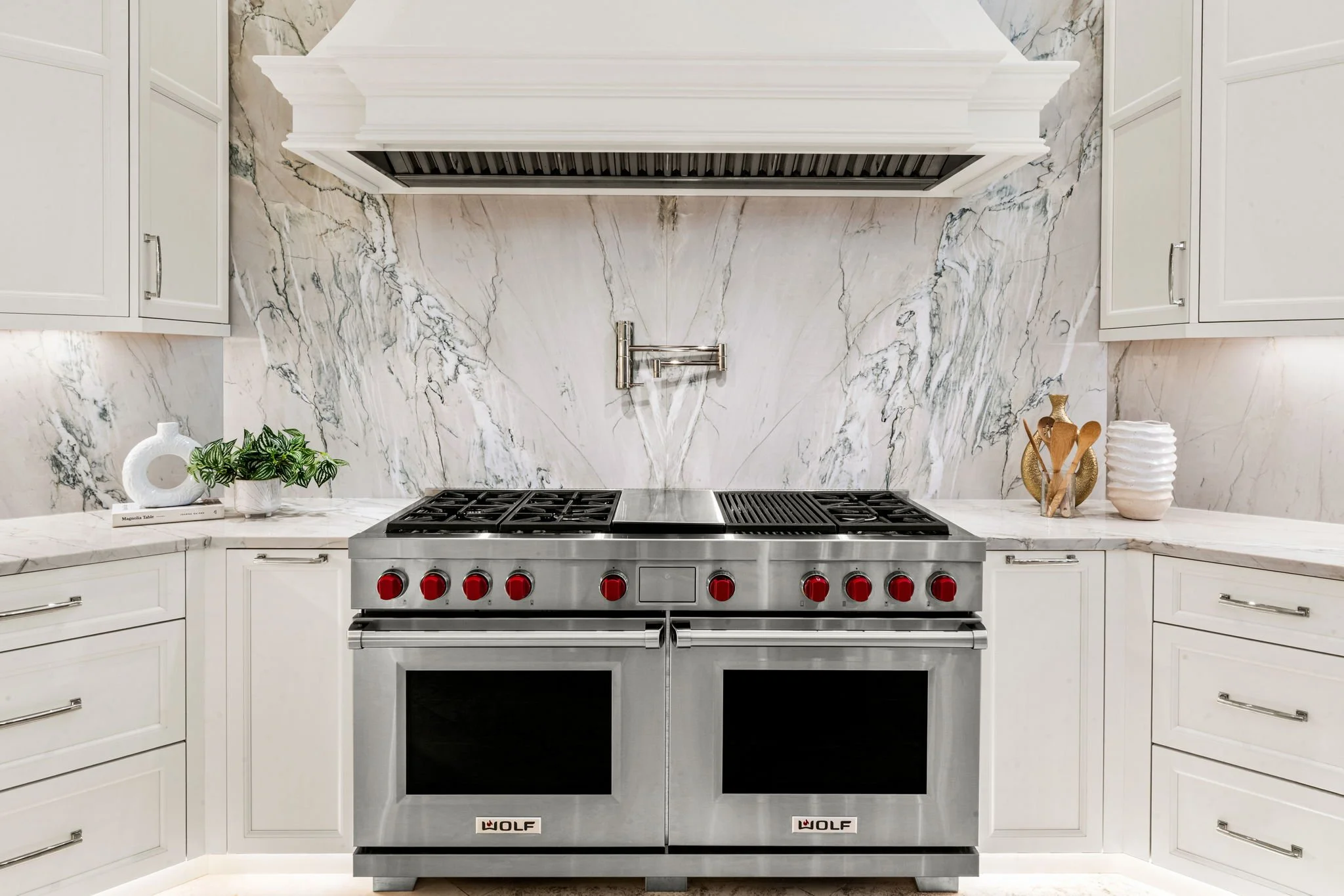 A modern kitchen with a stainless steel Wolf range stove, white cabinetry, marble backsplash, and some decorative items including a plant, vases, and bowls.