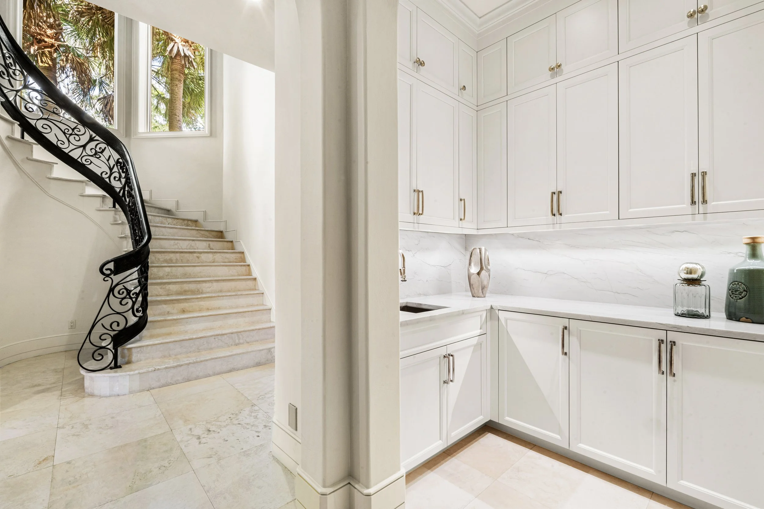 A spiral staircase with a black wrought iron railing next to a window with palm trees visible outside, and a white kitchen with marble countertops and white cabinets.