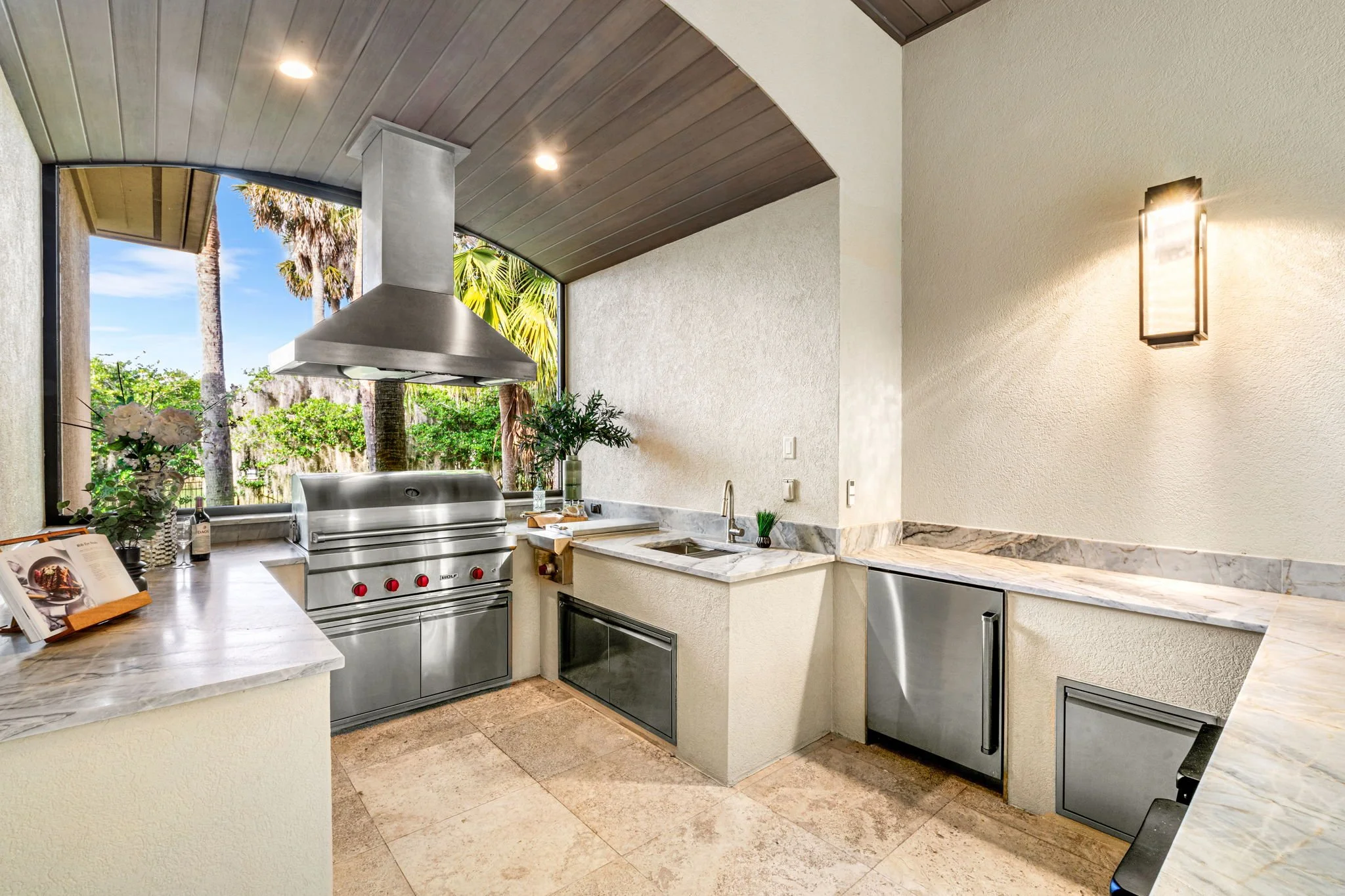 Outdoor kitchen with stainless steel grill, marble countertops, and a view of palm trees through a large window.