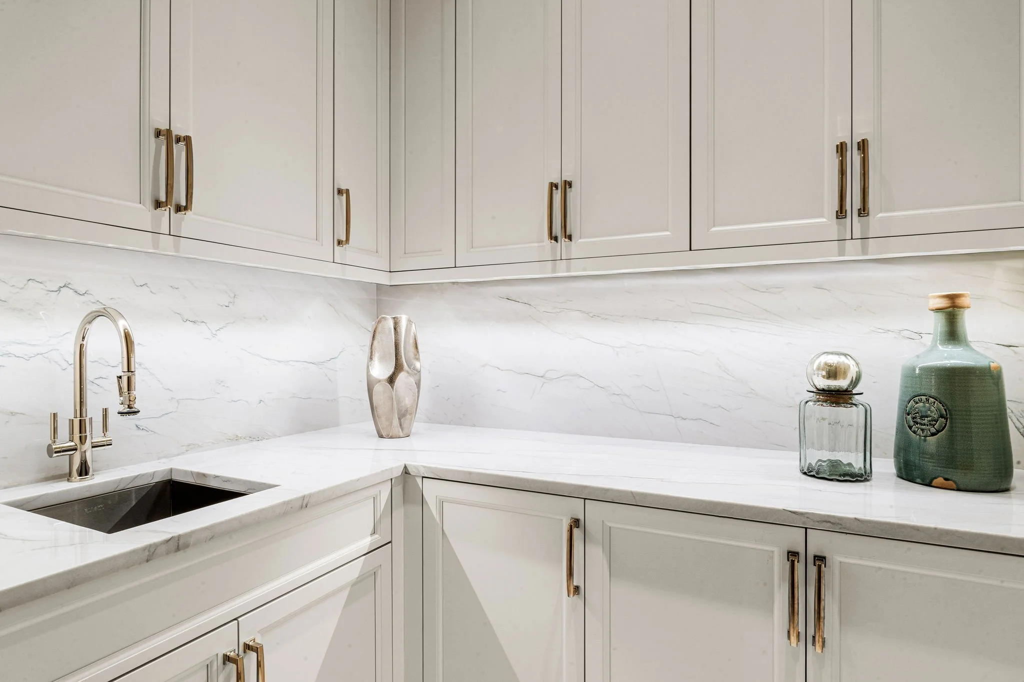 White kitchen with marble backsplash and countertop, gold cabinet handles, and decorative vases on the counter.