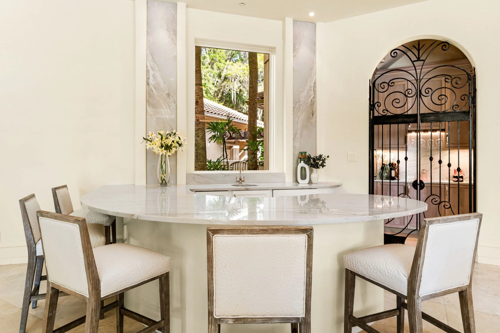 Modern kitchen with a round marble island, six upholstered chairs, a window showing trees outside, and a decorative wrought iron gate leading to another room.