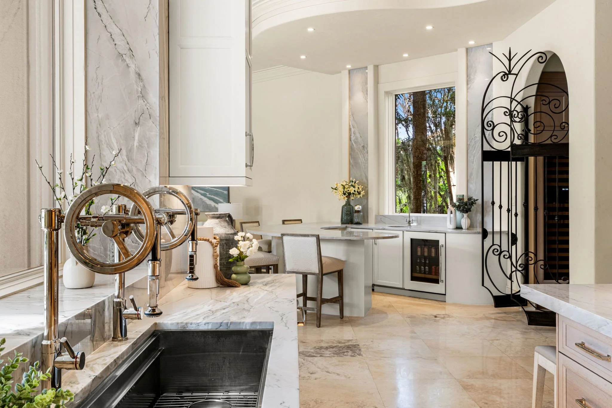 Modern kitchen with white marble countertops and a black undermount sink. Decor includes vases with white flowers, a small potted plant, and kitchen utensils. Large window with trees outside, recessed ceiling lights, and a spiral staircase with intricate black metalwork.