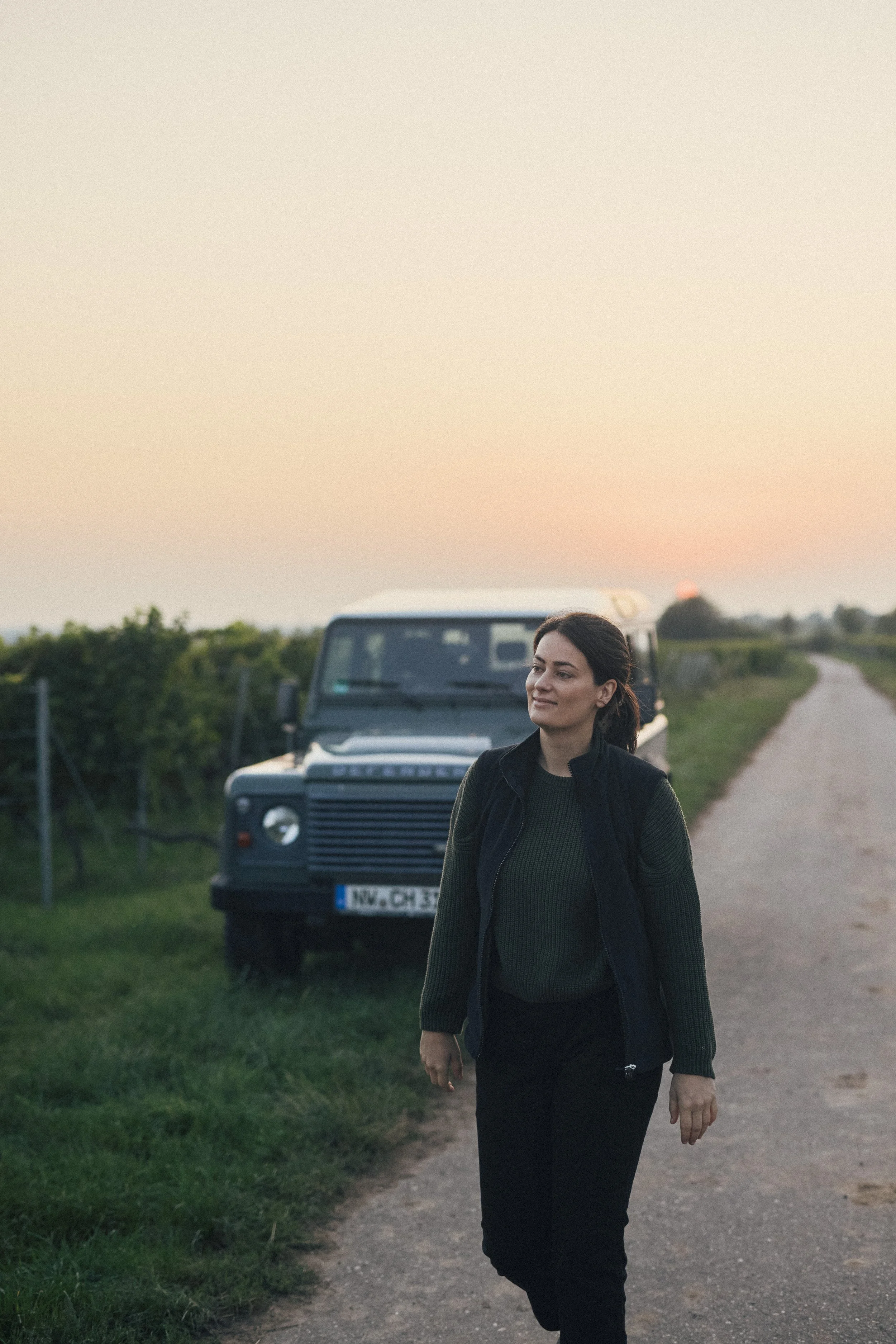 Frau läuft auf einem einer Landstraße neben einem schwarzen Land Rover bei Sonnenuntergang, mit grünem Feld und bewölktem Himmel im Hintergrund.