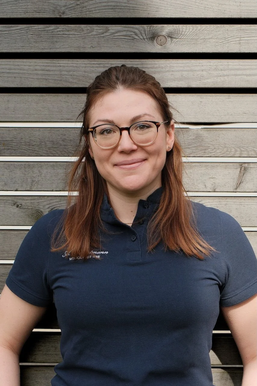 Porträt einer lachenden Frau mit roten Haaren, Brille und blauer Polo-Hemd, stehend vor einer Holzverkleidung.